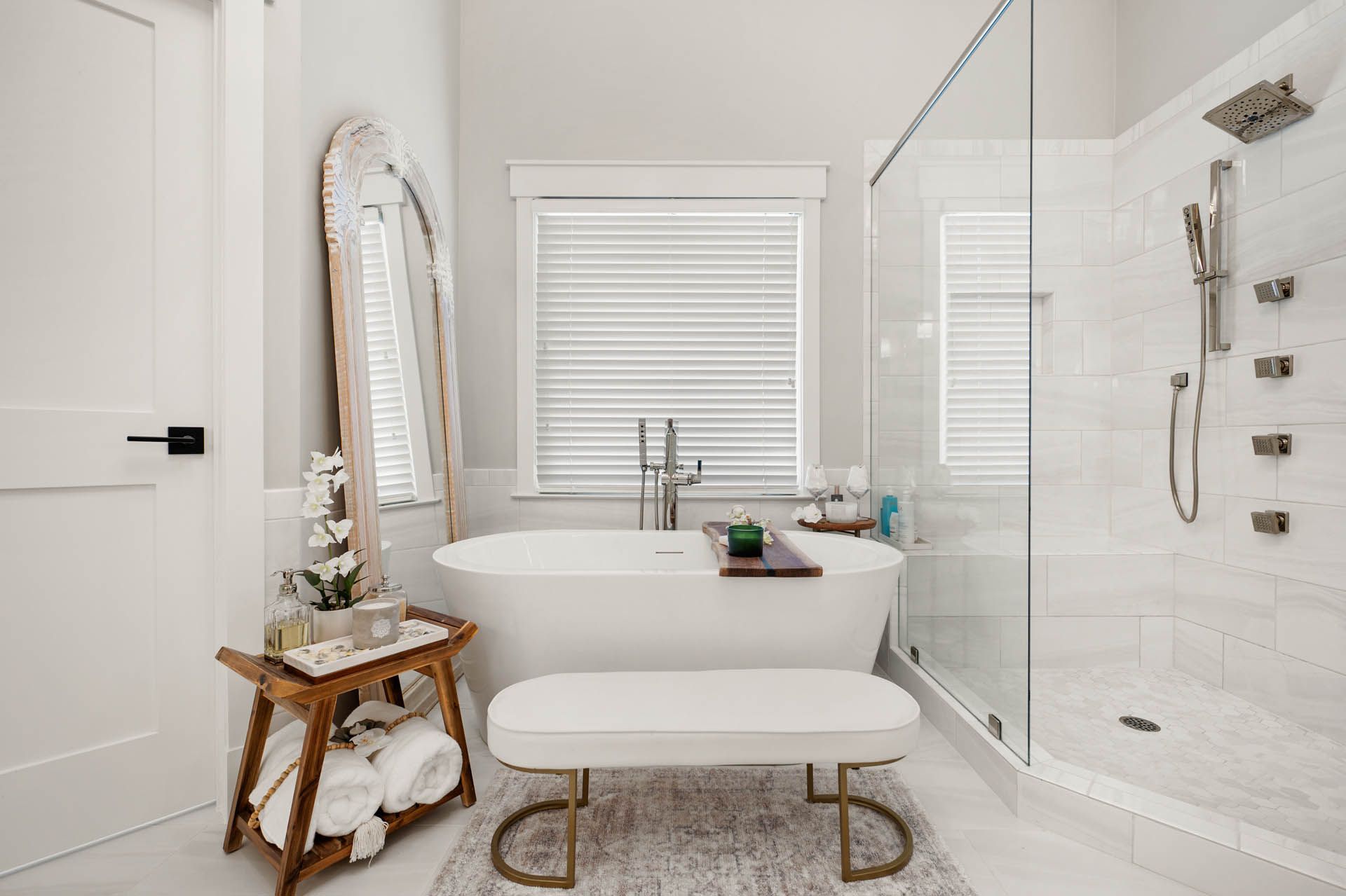 A bright white bathroom with a free-standing tub, glass shower, and wooden stool with towels.