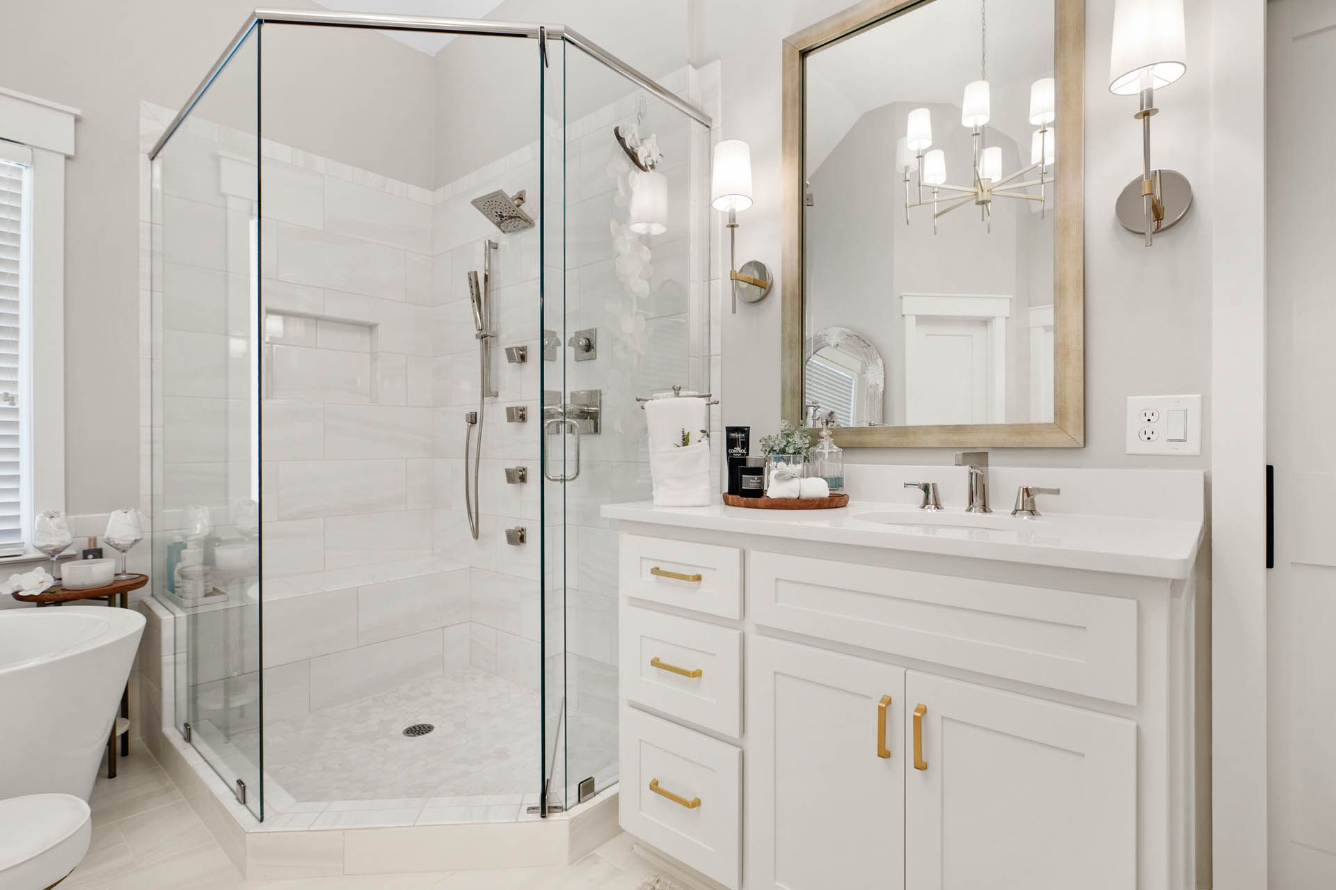Bright white bathroom with a glass-enclosed shower, vanity, and gold accents. A large mirror and chandelier hang above the sink.