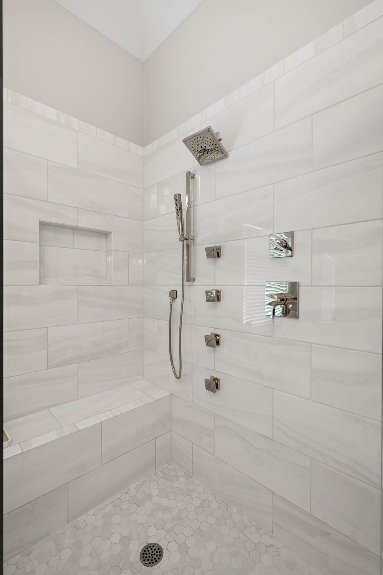 A modern, tiled shower with a built-in bench. Features a square showerhead, handheld sprayer, and multiple water controls on the wall.