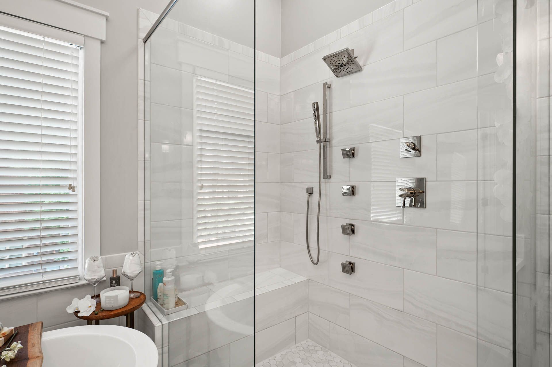 A modern bathroom with a walk-in shower featuring white tile, glass doors, and a rainfall showerhead.  A window with white blinds is visible.
