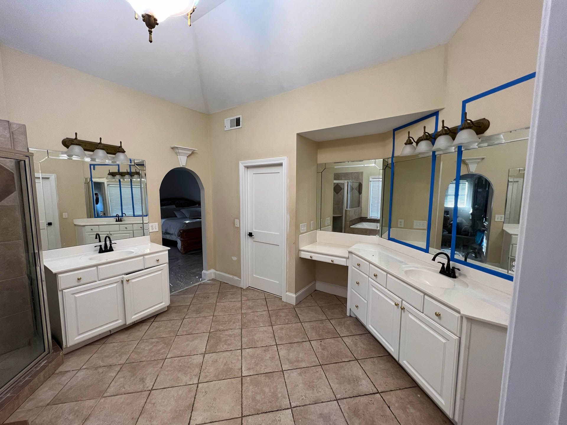 Spacious bathroom with dual vanities, beige tile floor, and large mirrors. A door leads to a bedroom and a shower is visible on the left.