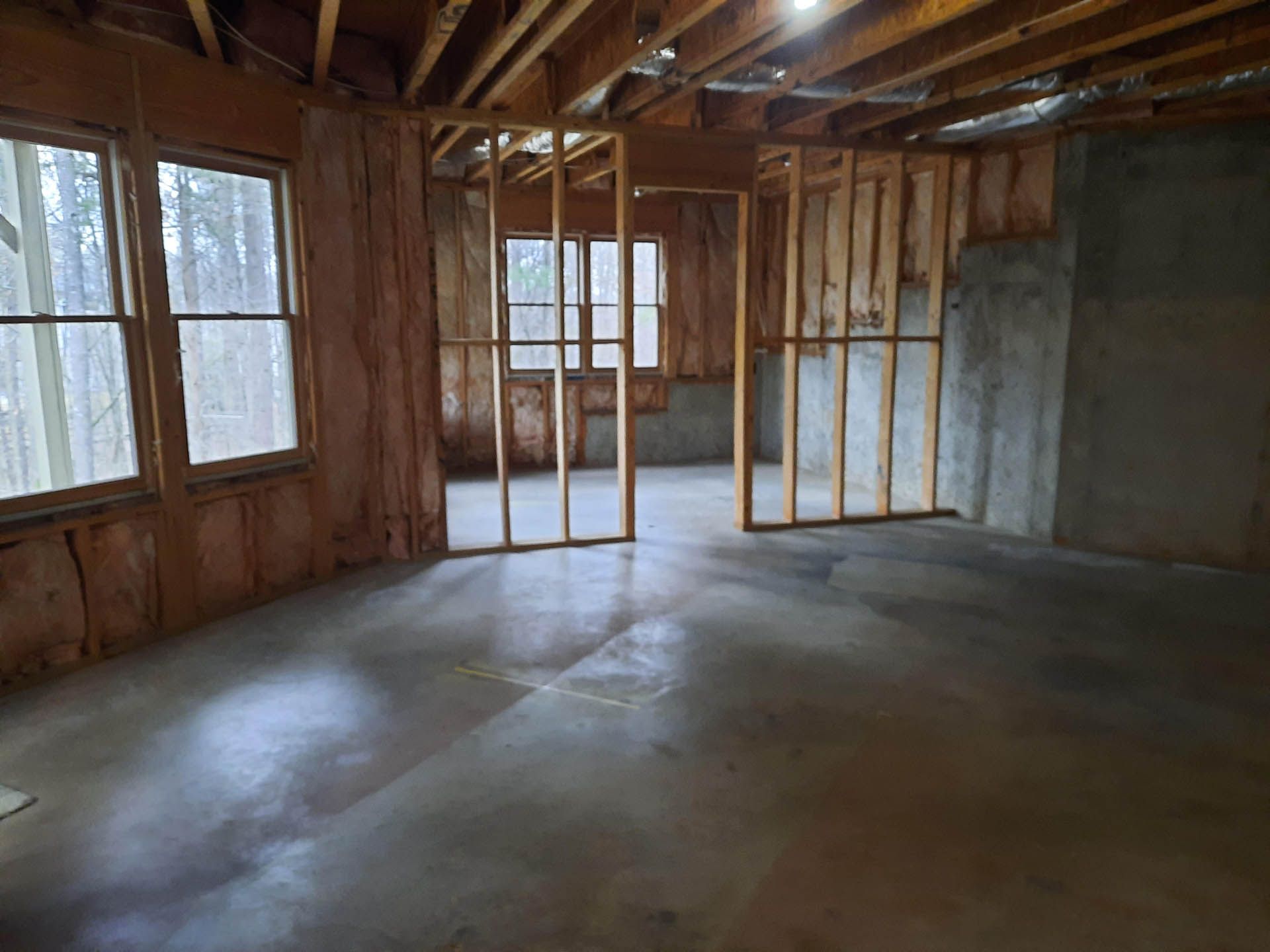 Unfinished room interior with concrete floor, wood-framed walls, and windows. Studs and unfinished walls are visible.