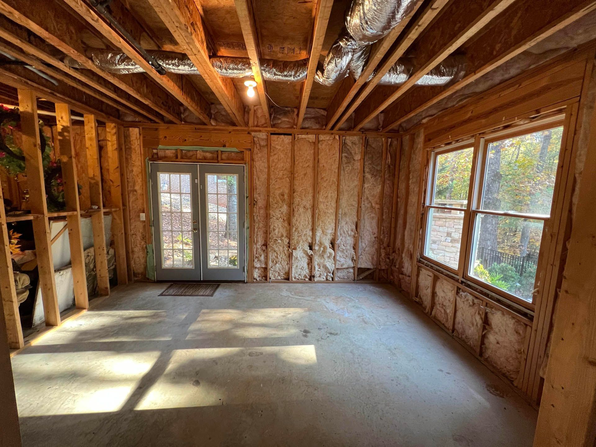 Unfinished room interior with bare wooden studs, exposed ceiling beams, and windows with sunlight.  French doors lead outside.