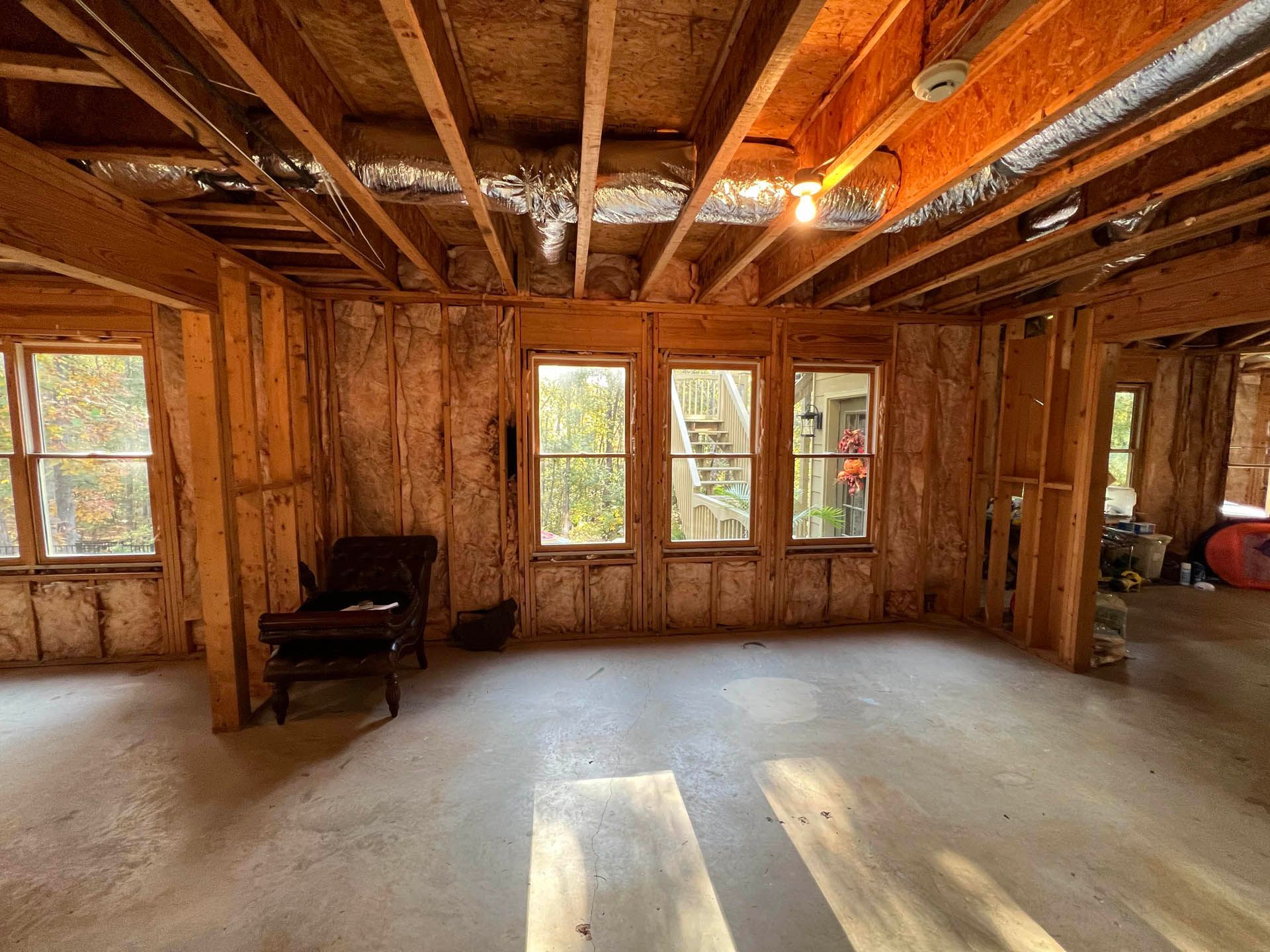 Interior of a room under construction with exposed wooden studs, insulation, windows, and a concrete floor. A chair sits in the room.