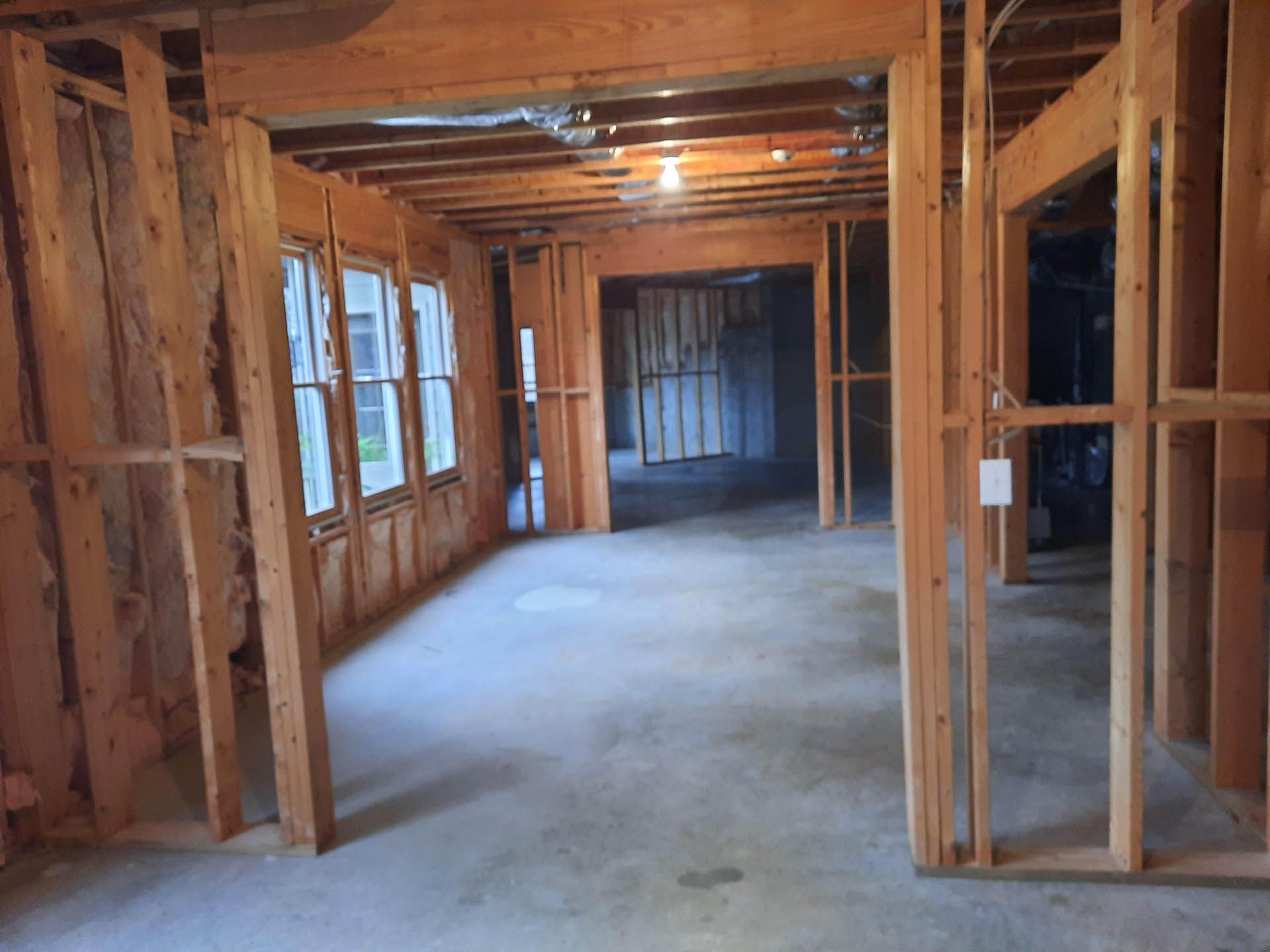 Interior view of a room under construction; exposed wooden framing and concrete floor. Natural light streams through a window, and the room has open doorways.