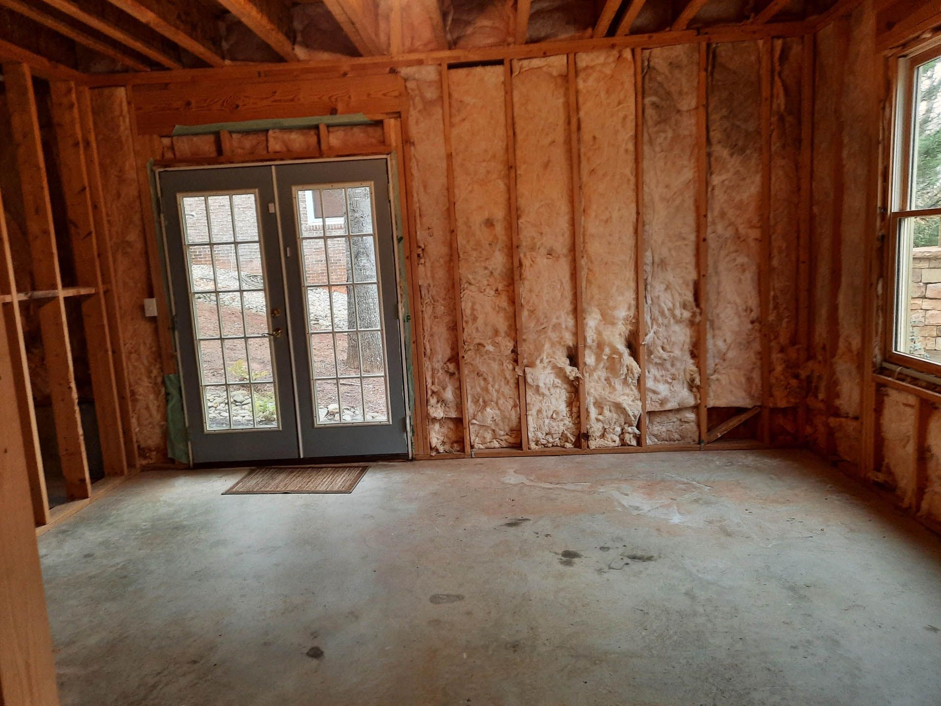 Interior of a room under construction with exposed wooden studs, concrete floor, and a double-door leading outside.