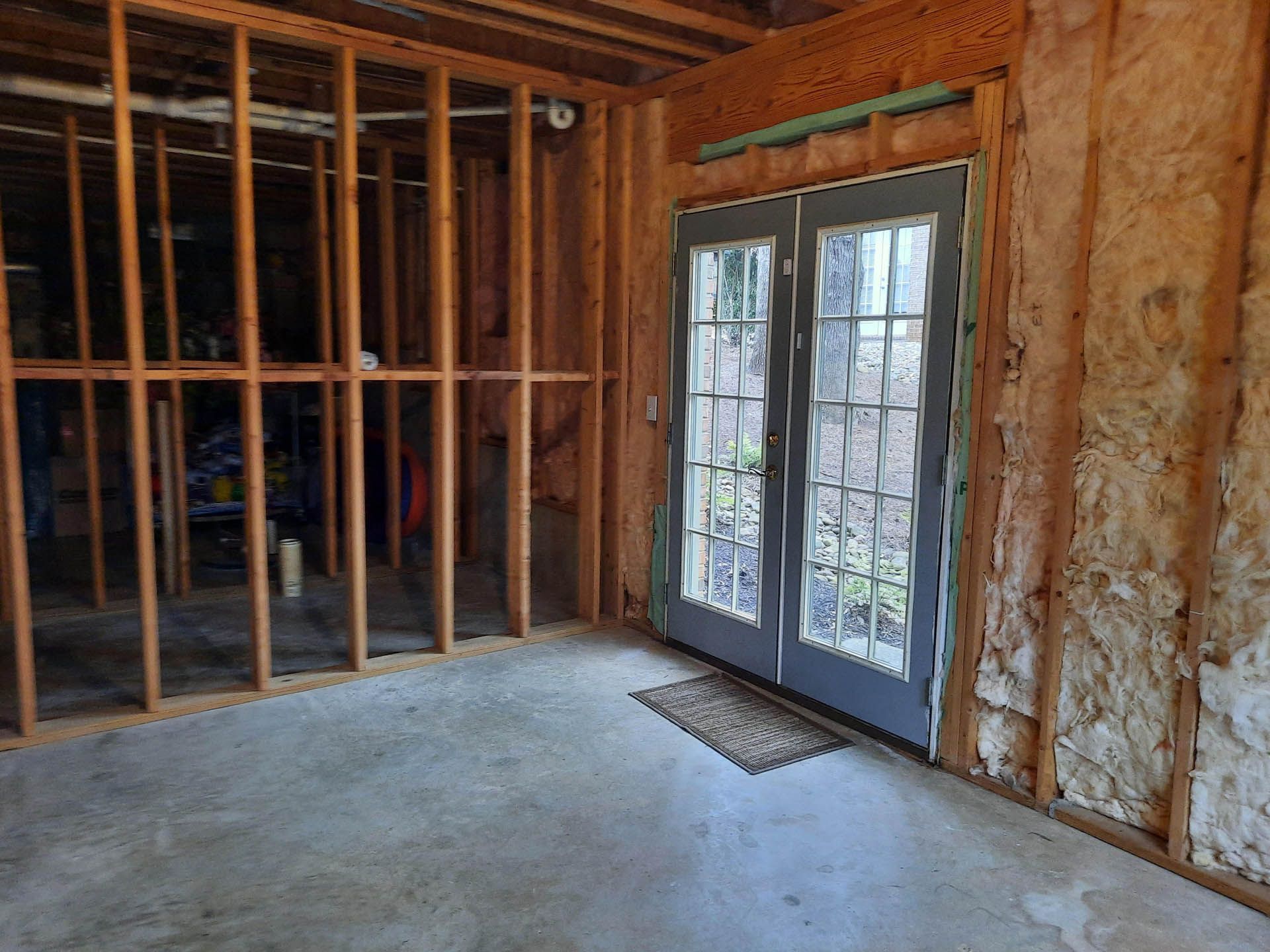 Interior view of a room under construction with unfinished walls, framing, and a double door leading outside. Concrete floor and insulation are visible.