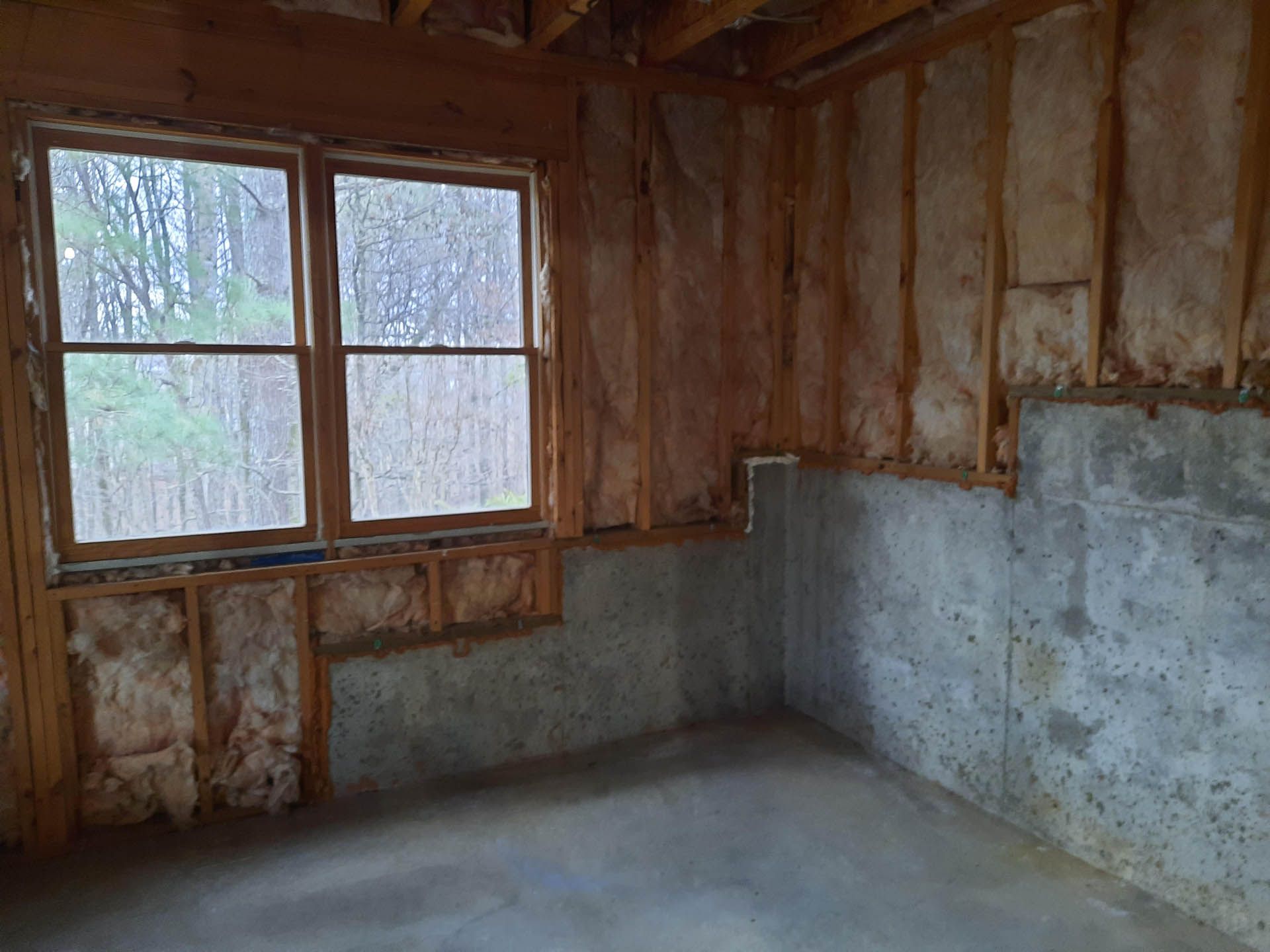 Interior of an unfinished room with a concrete foundation and wooden frame. A window and insulation are visible.