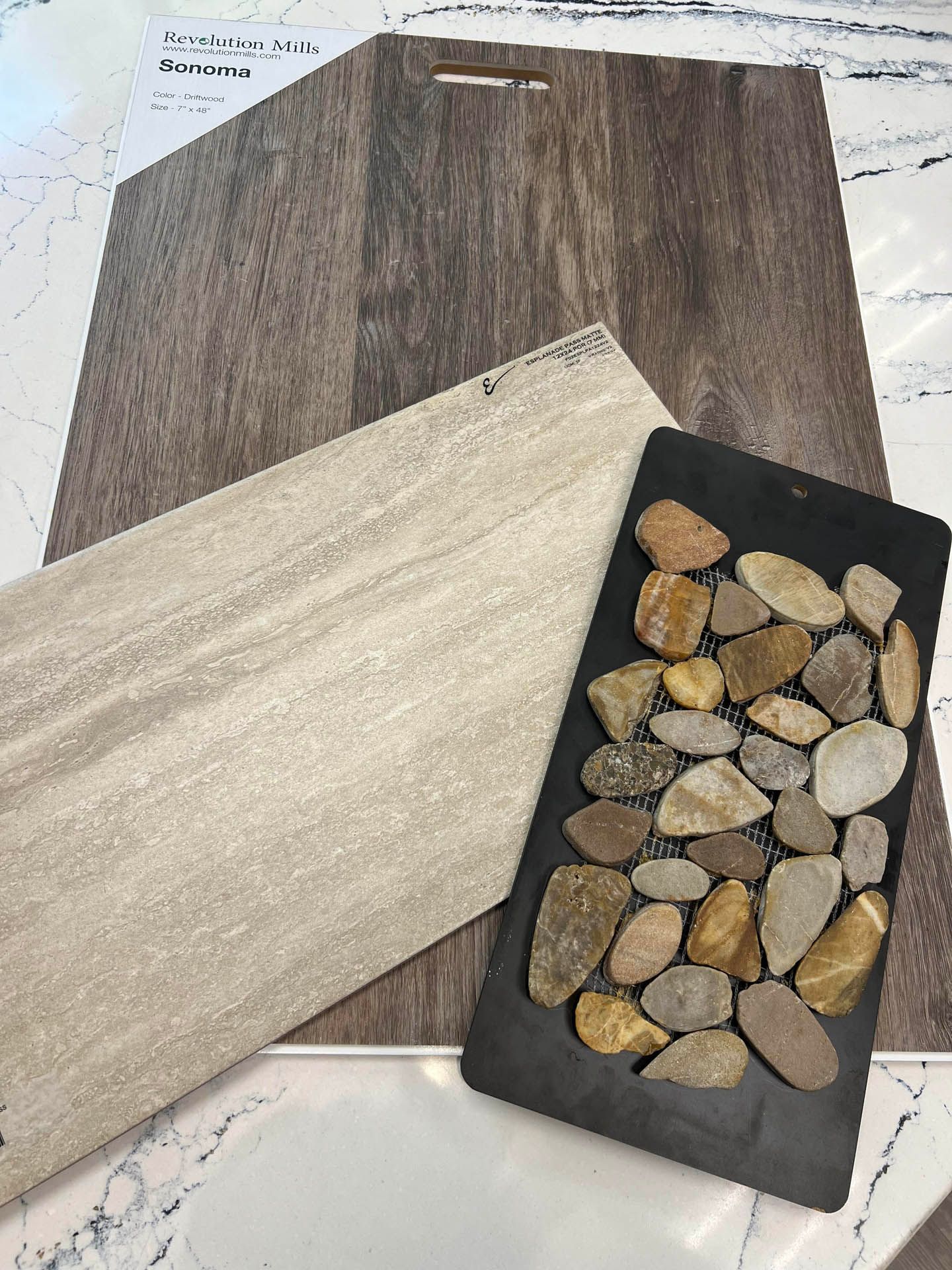 Samples of flooring and decorative tile on a marble countertop: wood-look, light gray, and river rock.