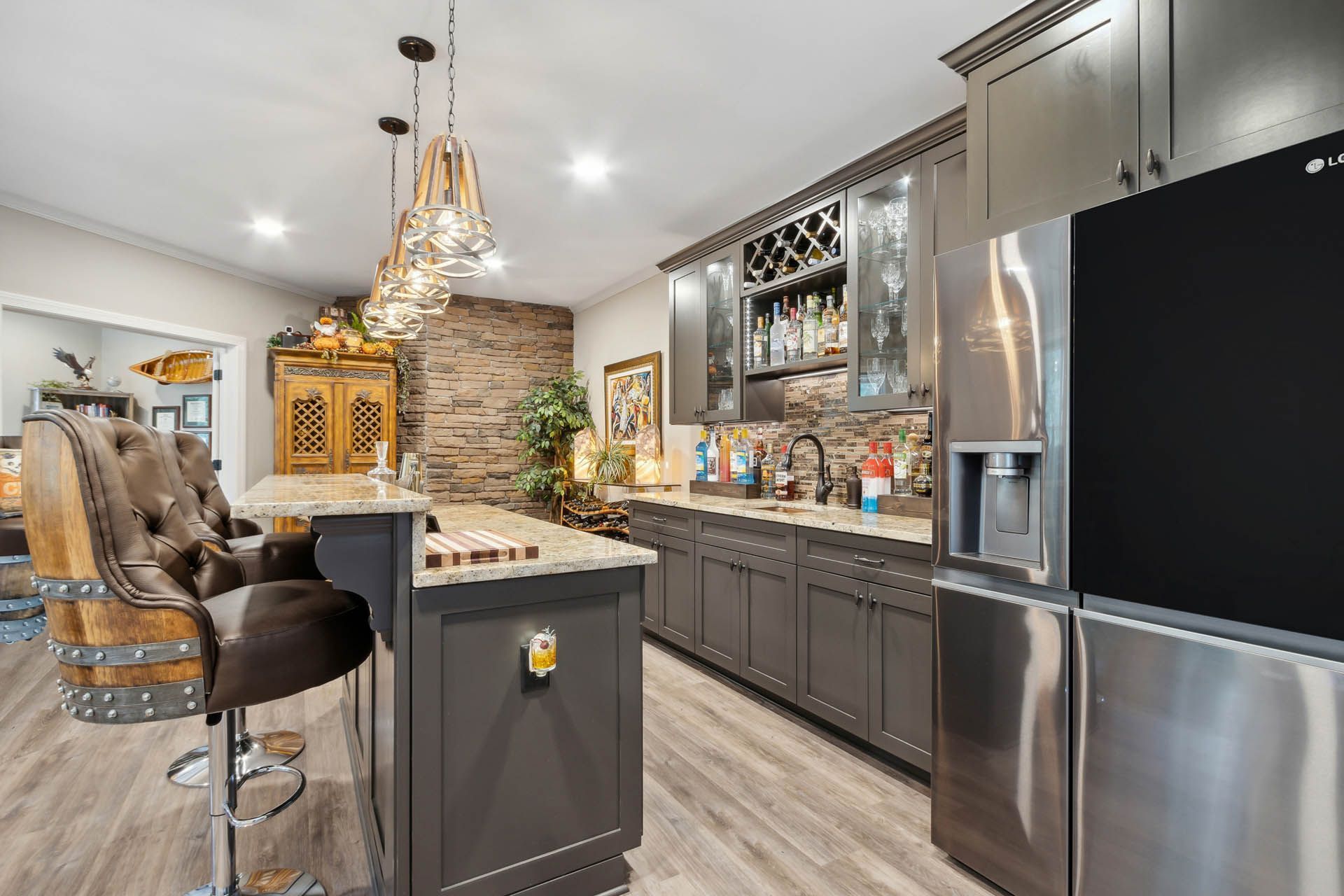 A stylish bar area with dark gray cabinets, a stone backsplash, and a stainless steel refrigerator. Two cushioned bar stools sit at the island.