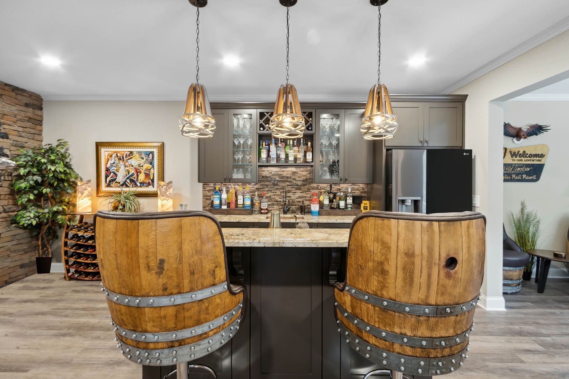 A home bar with barrel-shaped stools, overhead pendant lights, and liquor bottles on display.  A refrigerator and cabinet are also visible.