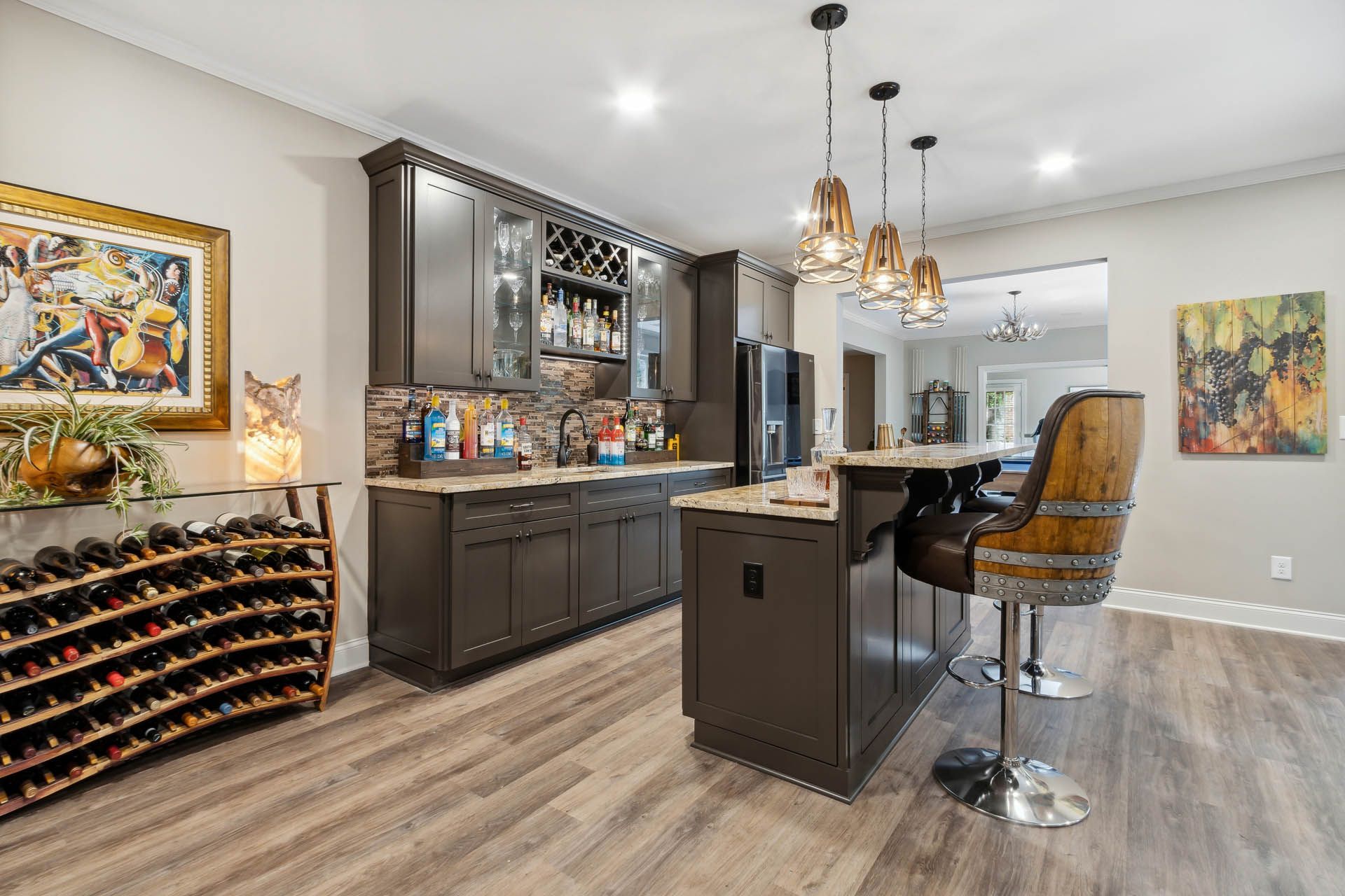 A home bar with dark cabinets, a countertop, and a small island with a bar stool. Wine racks and artwork are also visible.