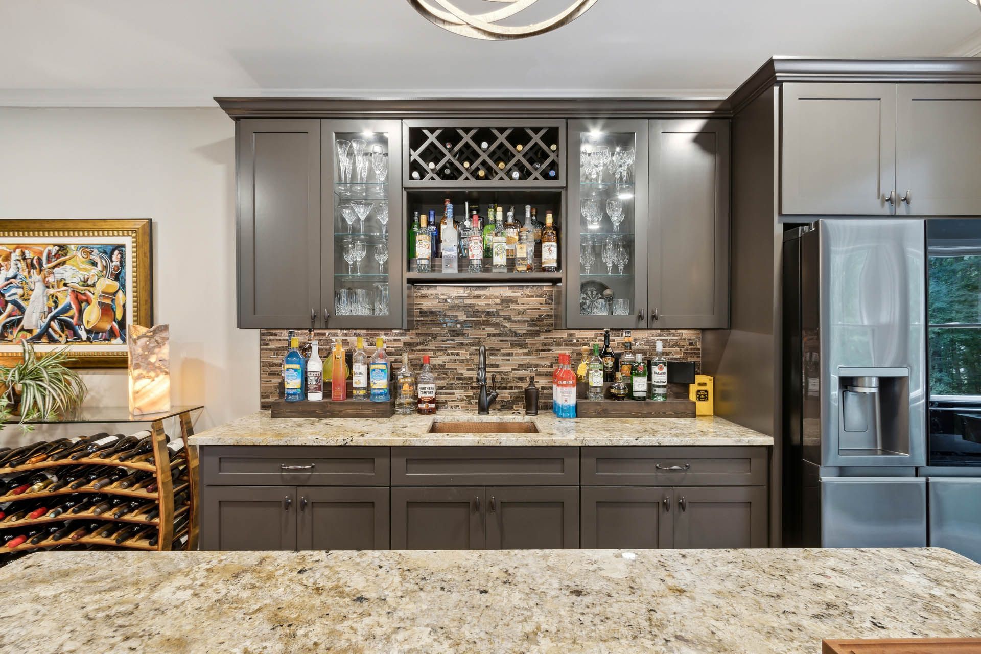 Home bar with granite countertop, dark cabinetry, glass-fronted display cabinets for liquor bottles, and a small sink. A painting and a refrigerator are nearby.