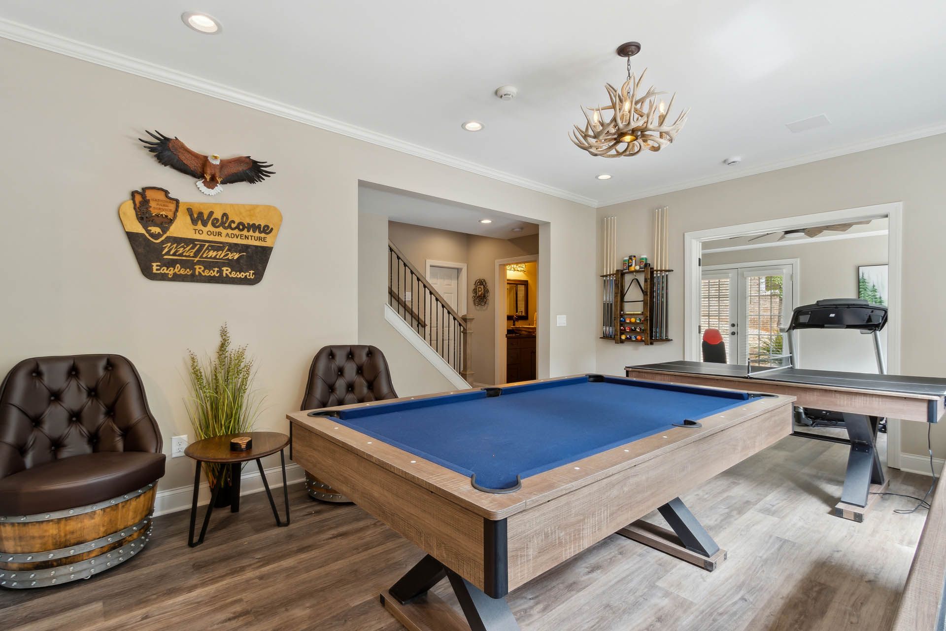 A recreation room with a pool table, chairs, and a treadmill. The room has a rustic feel with wooden elements and warm lighting.
