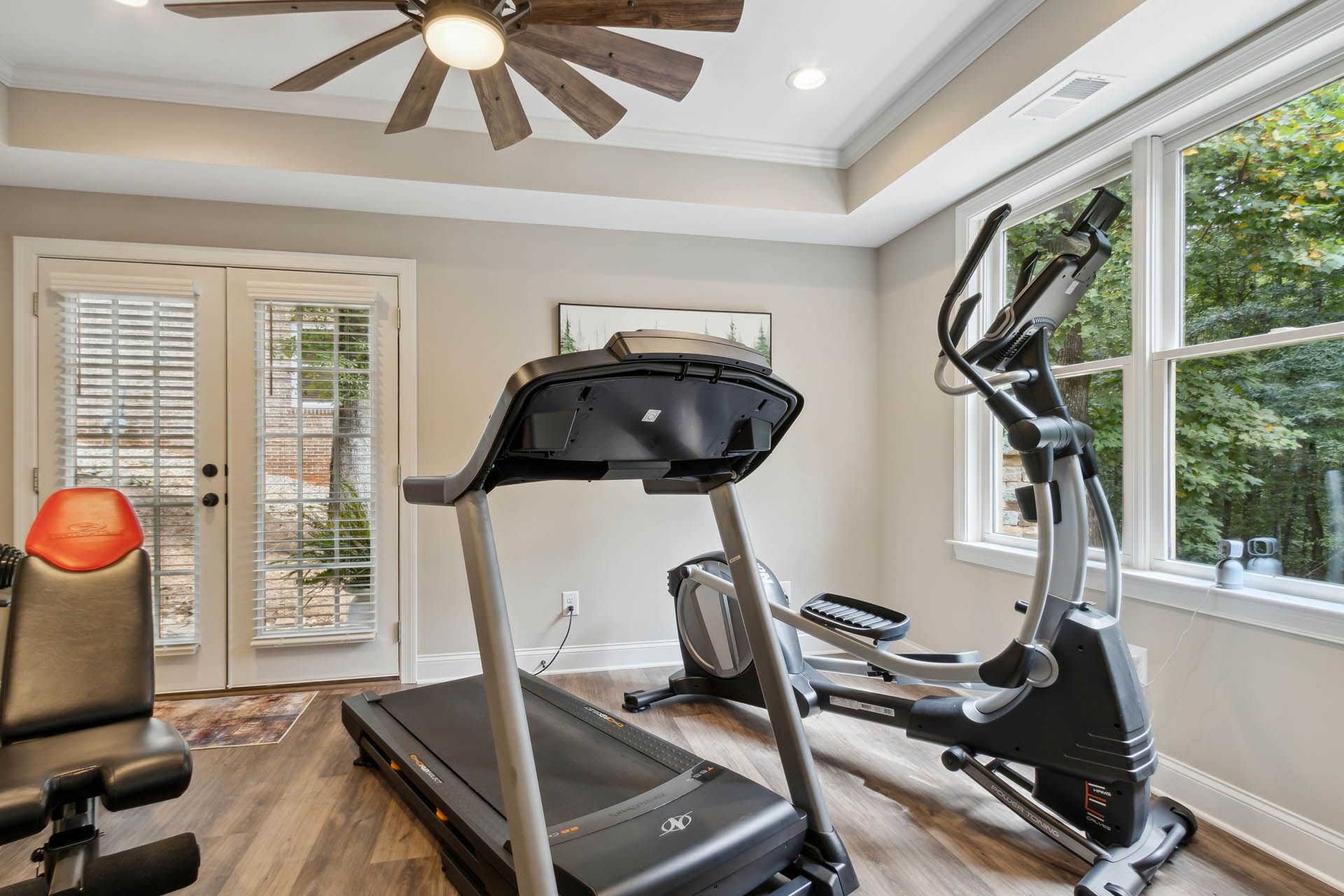 A home gym with a treadmill, elliptical machine, and weight bench. Sunlight streams in from a window and French doors.