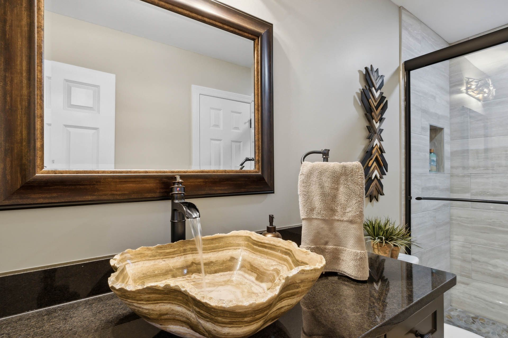 Bathroom with a decorative stone sink, black countertop, large mirror, and a shower with gray tiled walls.