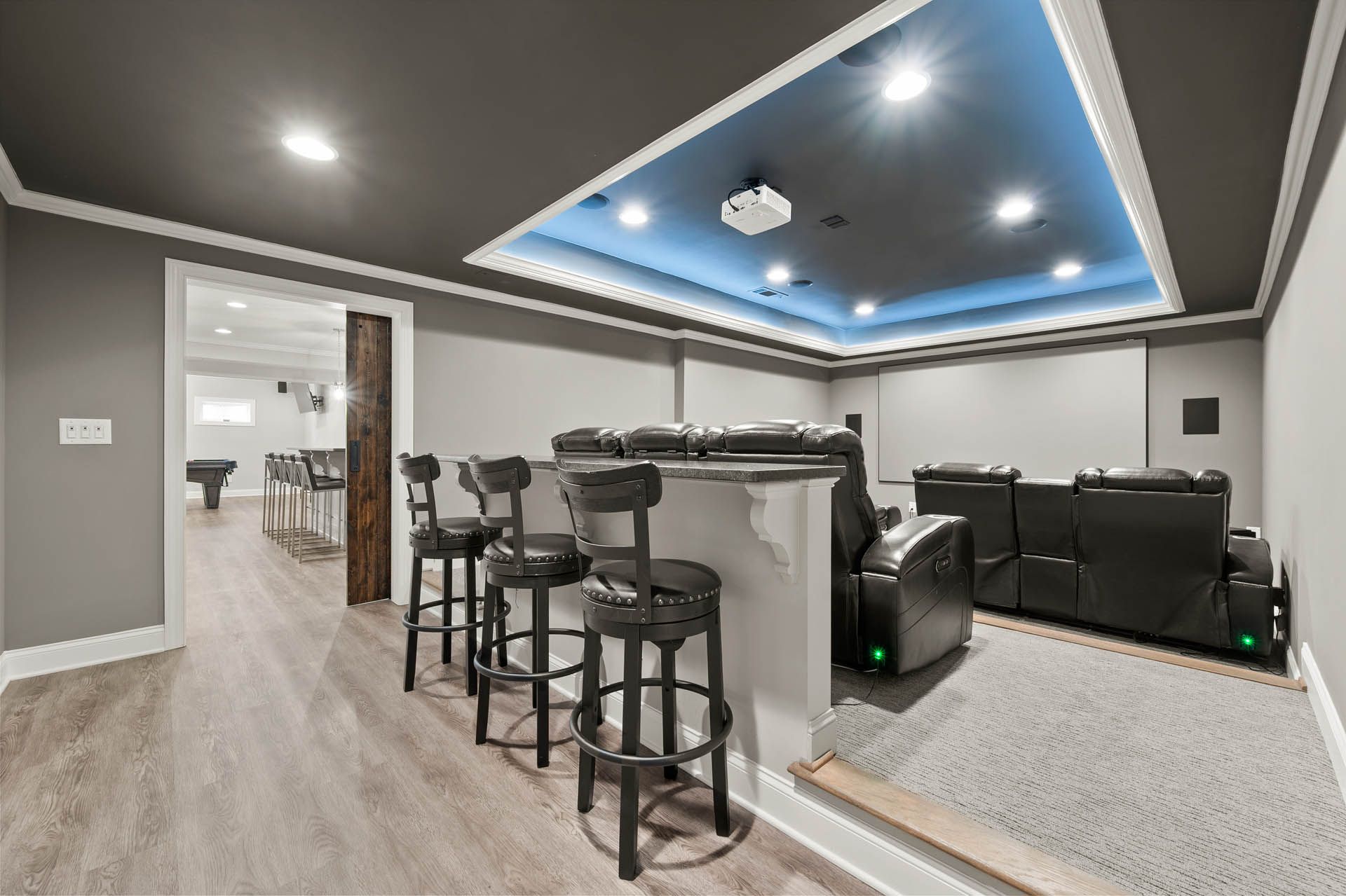 Home theater with a bar, grey walls, and blue ceiling lighting. A projection screen is visible, along with black leather seating and bar stools.