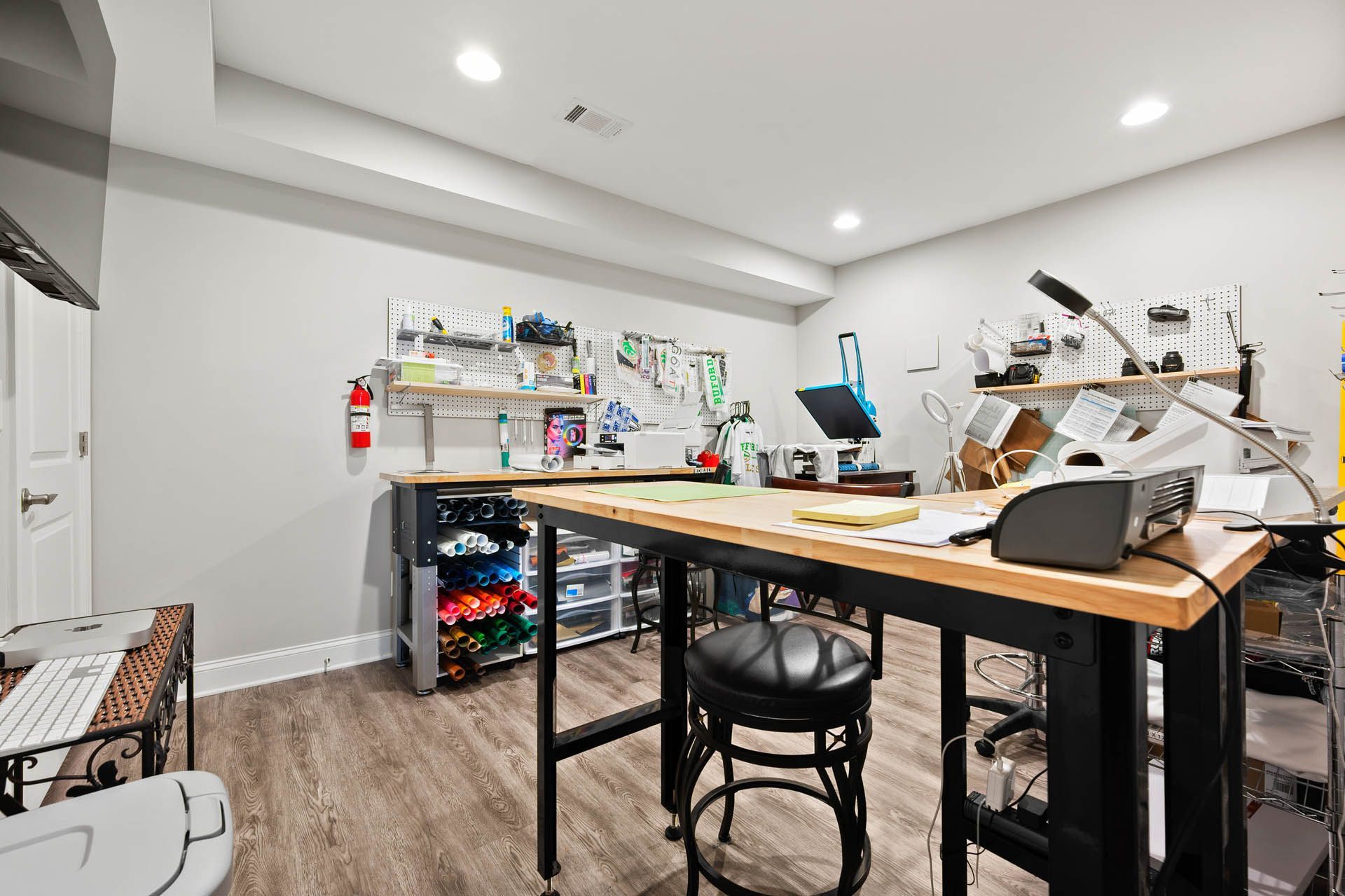 Workshop space with a large workbench, tools, and supplies. A fire extinguisher hangs on the wall near a door, and colorful items are stored on a shelf.