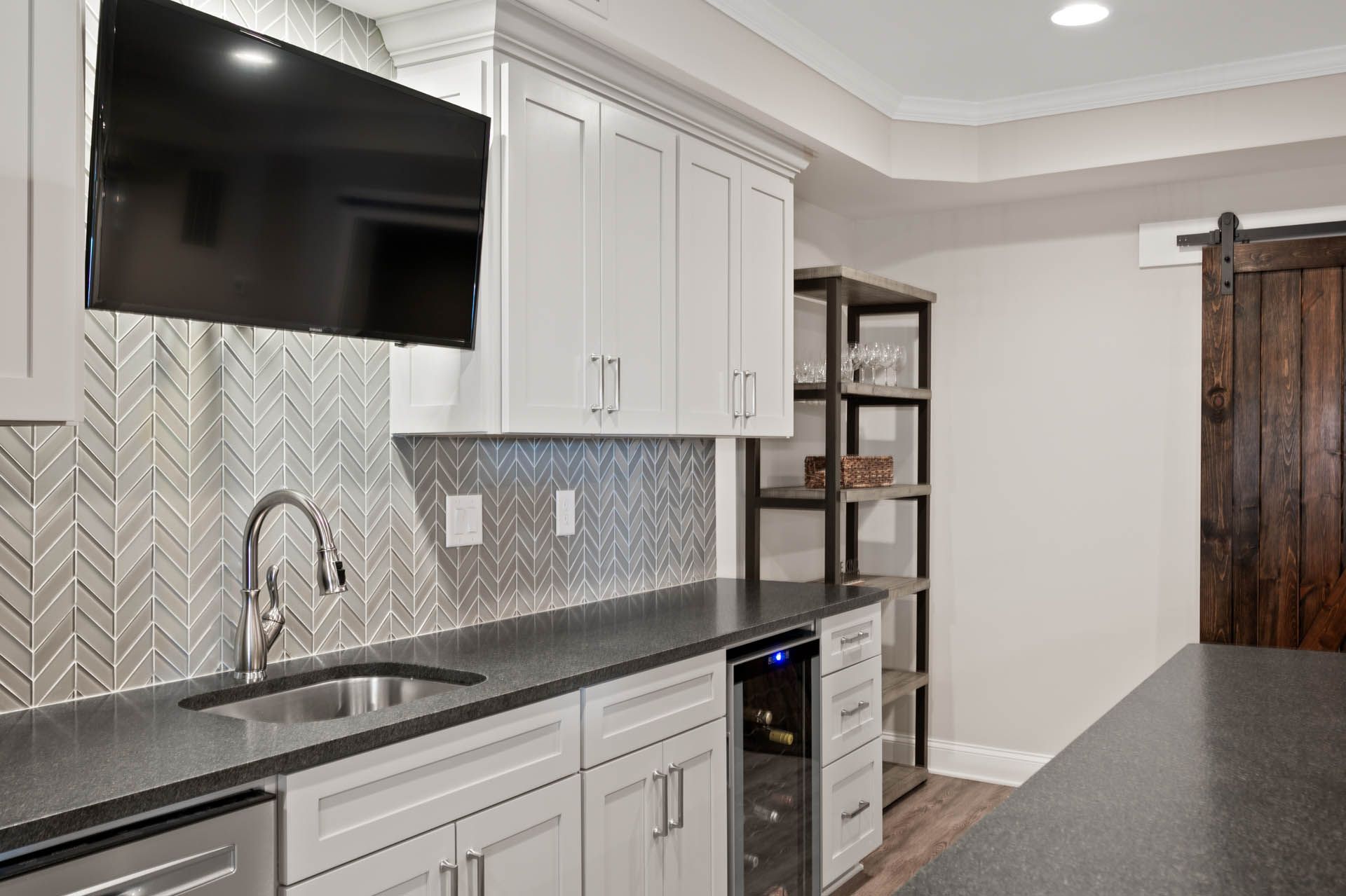 A basement wet bar with a TV, sink, cabinets, and wine cooler against a grey backsplash, next to a shelving unit and a dark wooden barn door.