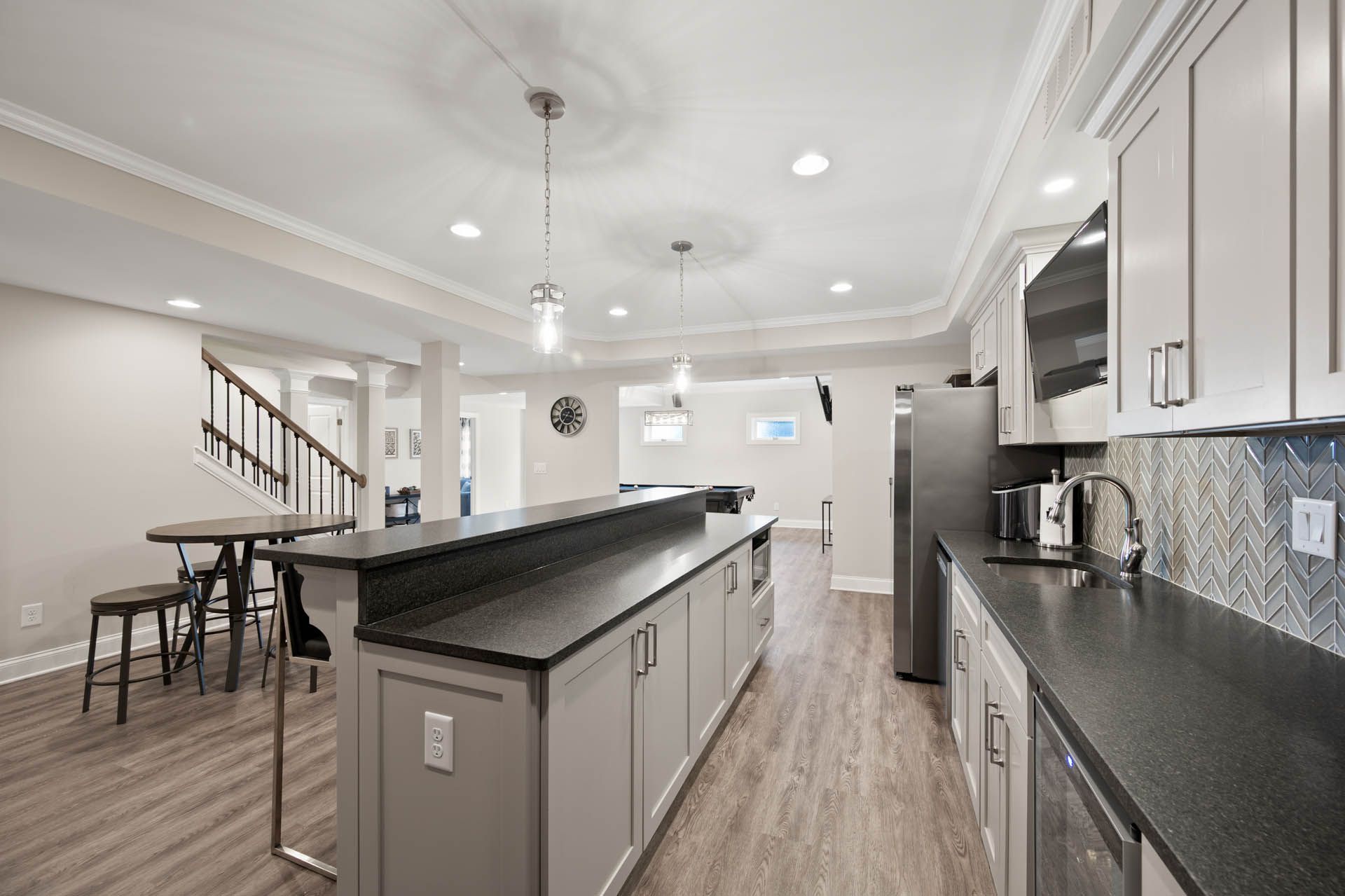 Modern kitchen with gray cabinets, dark countertops, and wood-look flooring. A large island with seating and a refrigerator are visible.