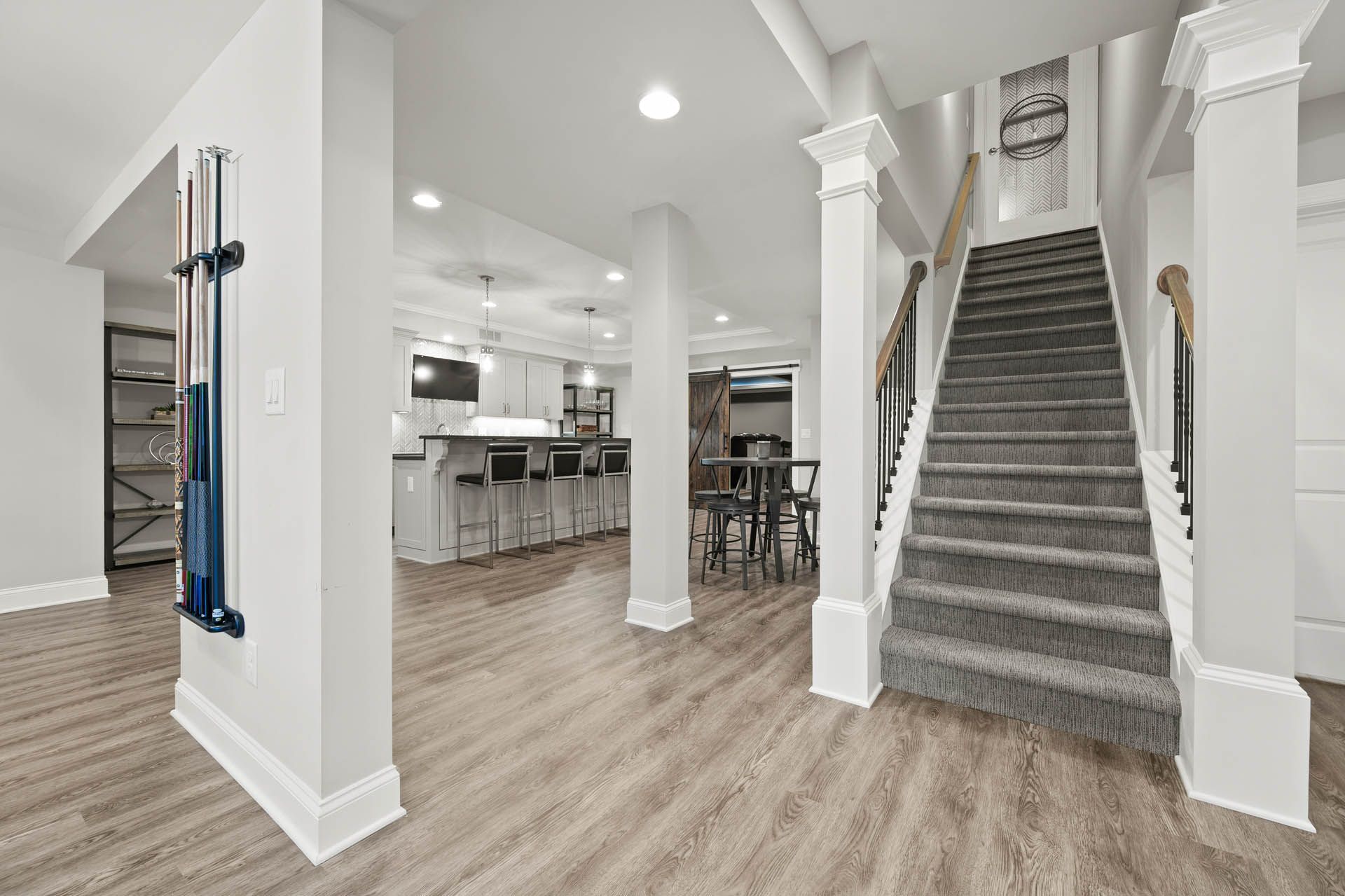 Basement with staircase, bar, and columns. Light wood-look flooring, white walls, and gray carpeted stairs.