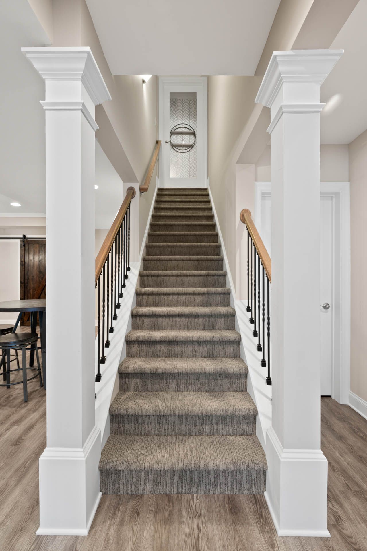 Staircase with beige carpet, flanked by white pillars and handrails, leading up to a door with a circular design element.
