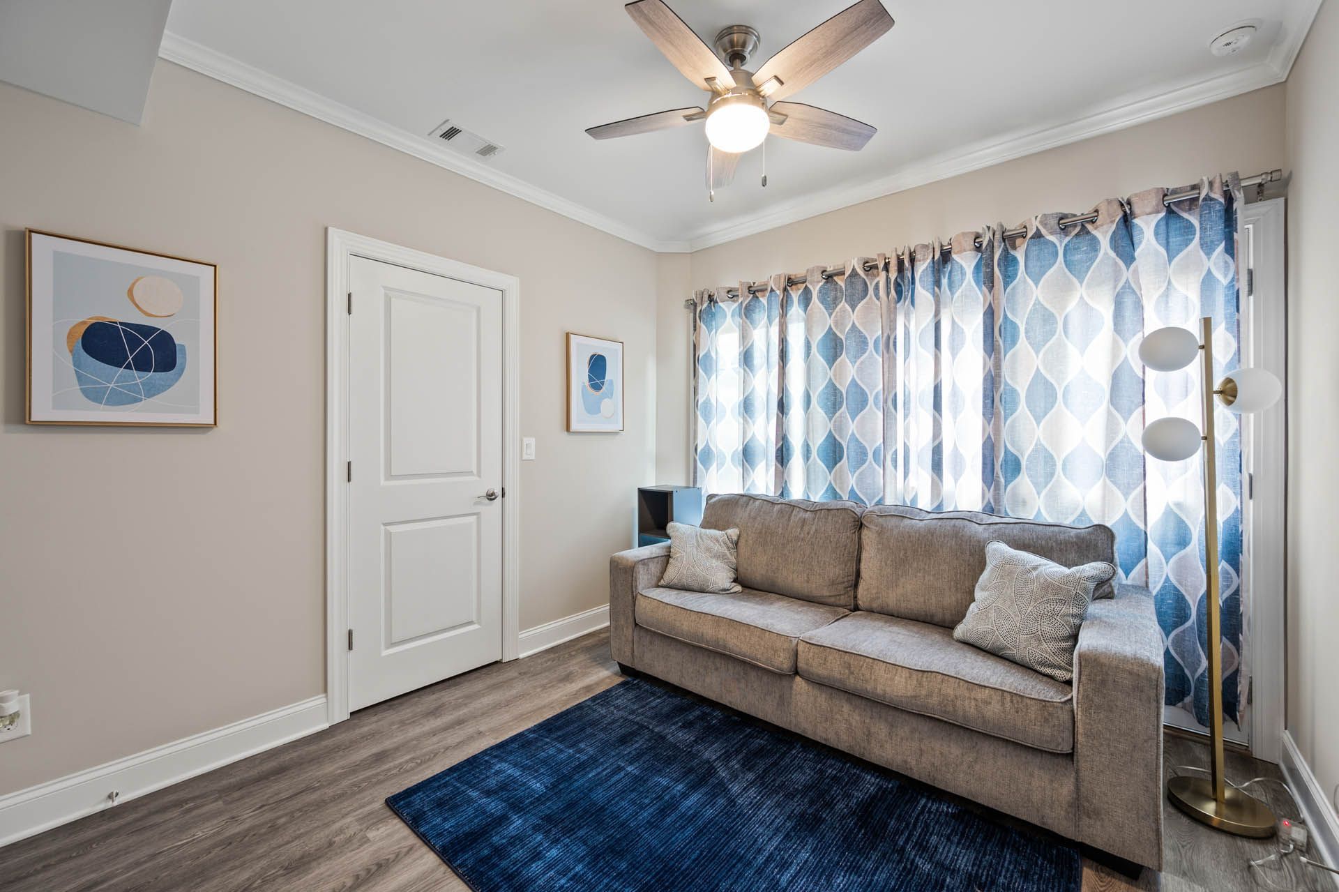 A small living area with a gray sofa, blue rug, patterned curtains, artwork, and a ceiling fan. A doorway is on the left.
