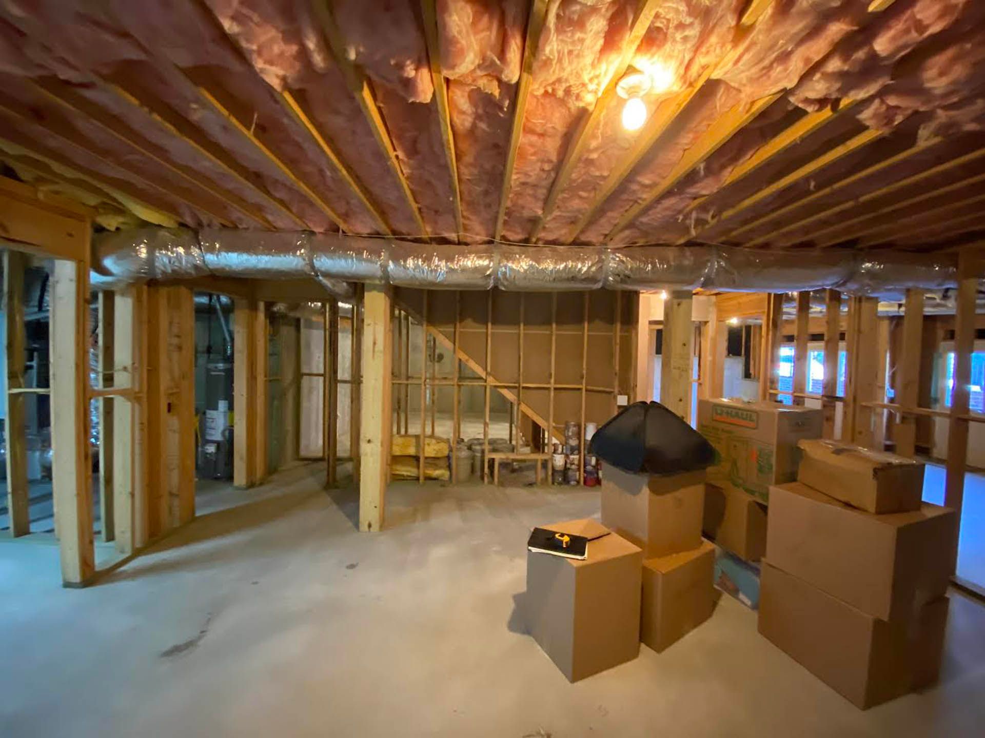 An unfinished basement with exposed wooden beams and insulation, HVAC ductwork, and cardboard boxes.