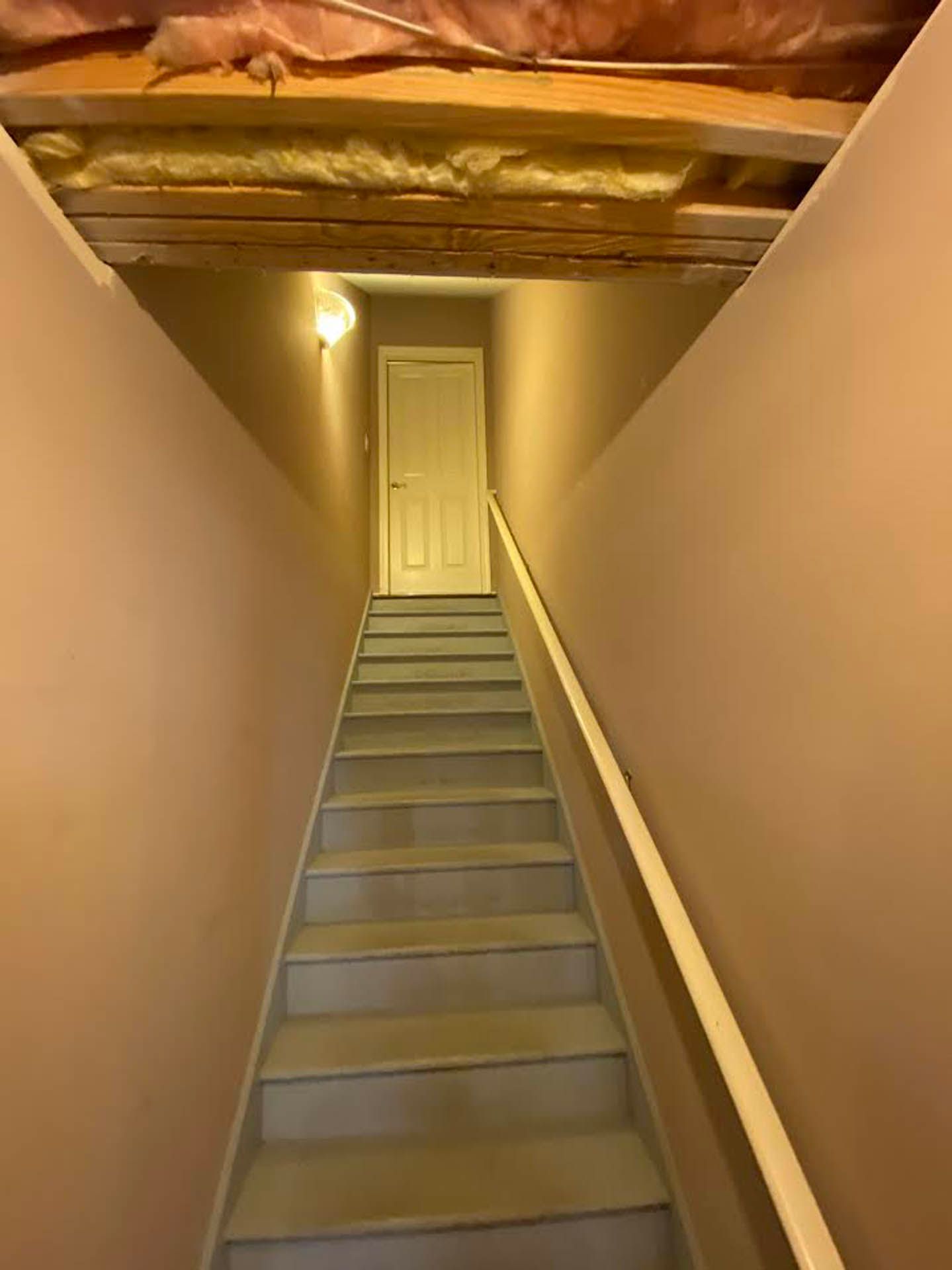 A narrow staircase leads up to a closed door. Beige walls and a wooden ceiling with exposed insulation frame the stairs.