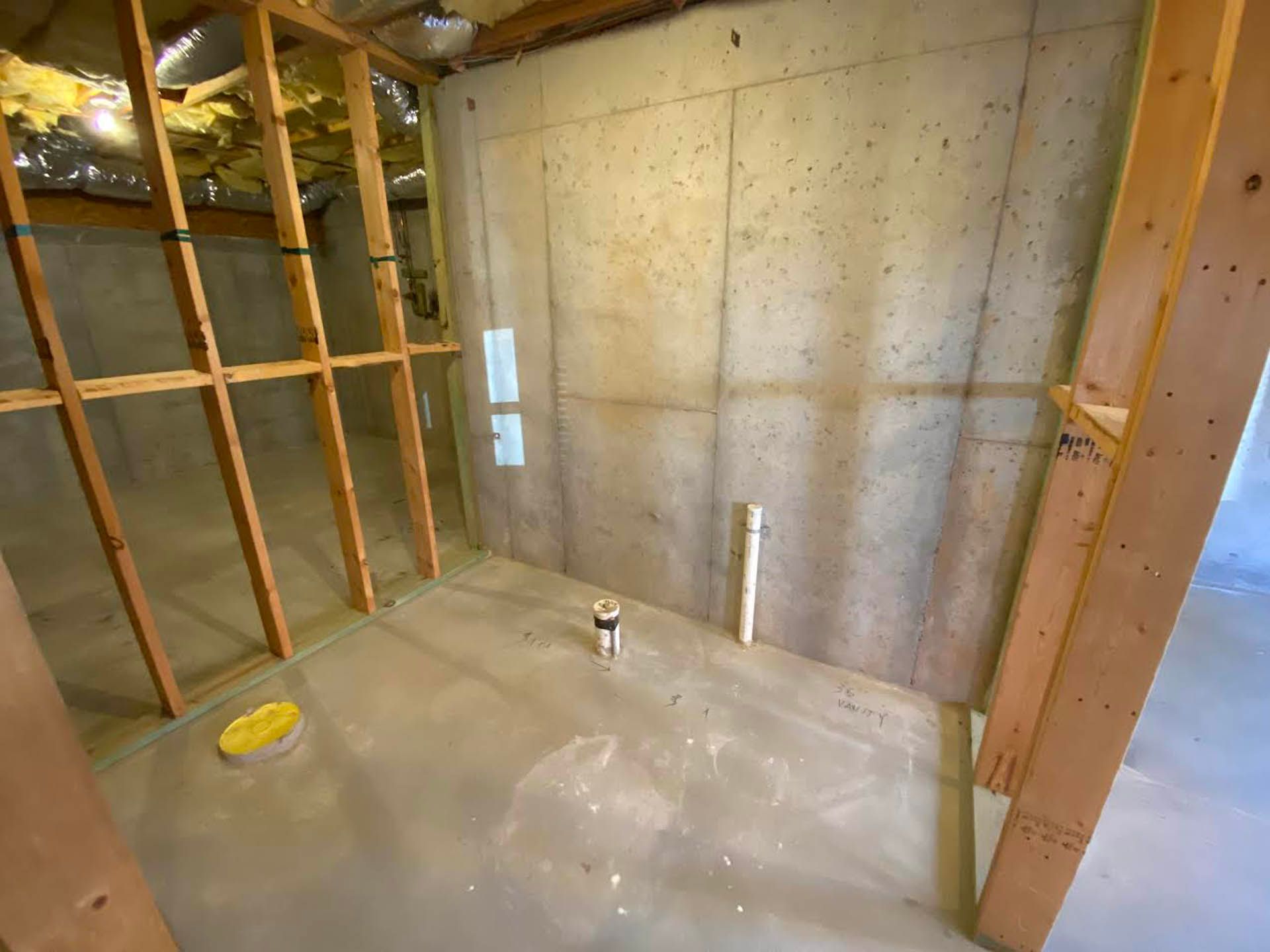 An unfinished basement room with concrete walls, wooden studs, and exposed plumbing. The room has a gray floor and a section with yellow insulation.