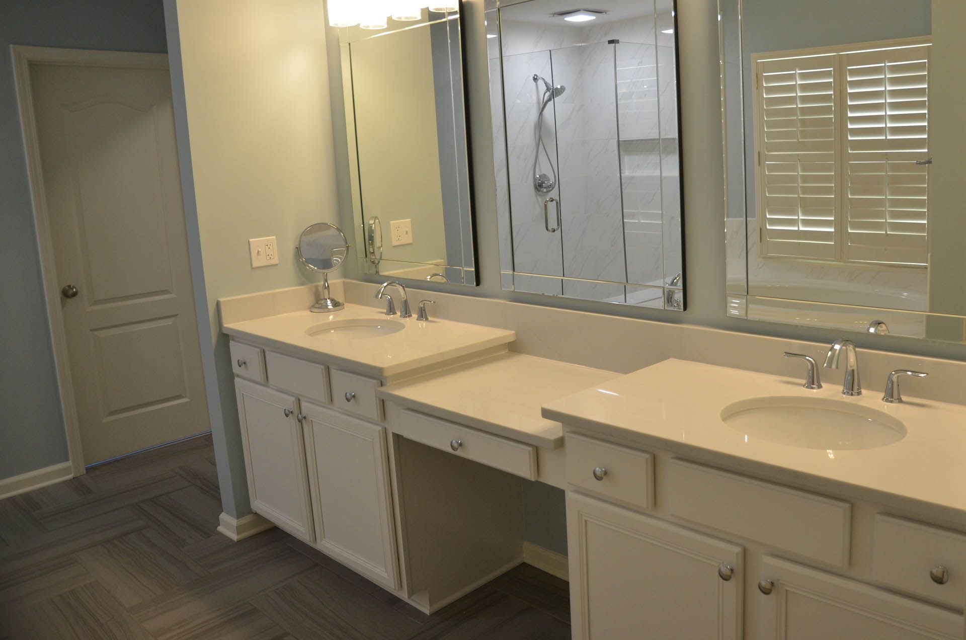 A modern bathroom with white countertops, two sinks, large mirrors, and a shower visible in the background. Blue walls and flooring.