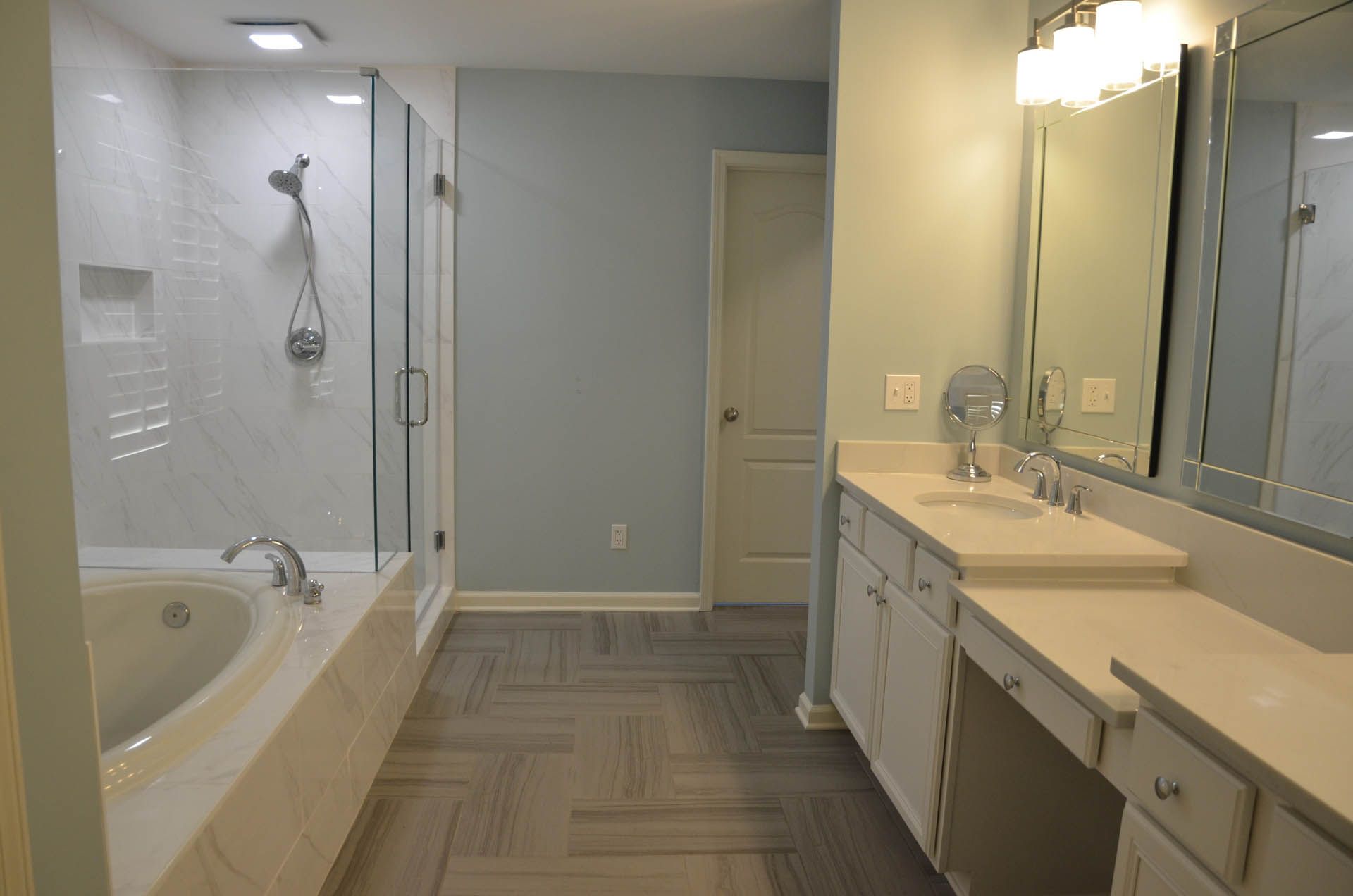 Spacious, light blue bathroom with a bathtub, glass-enclosed shower, and double sink vanity. Gray herringbone tile floor.