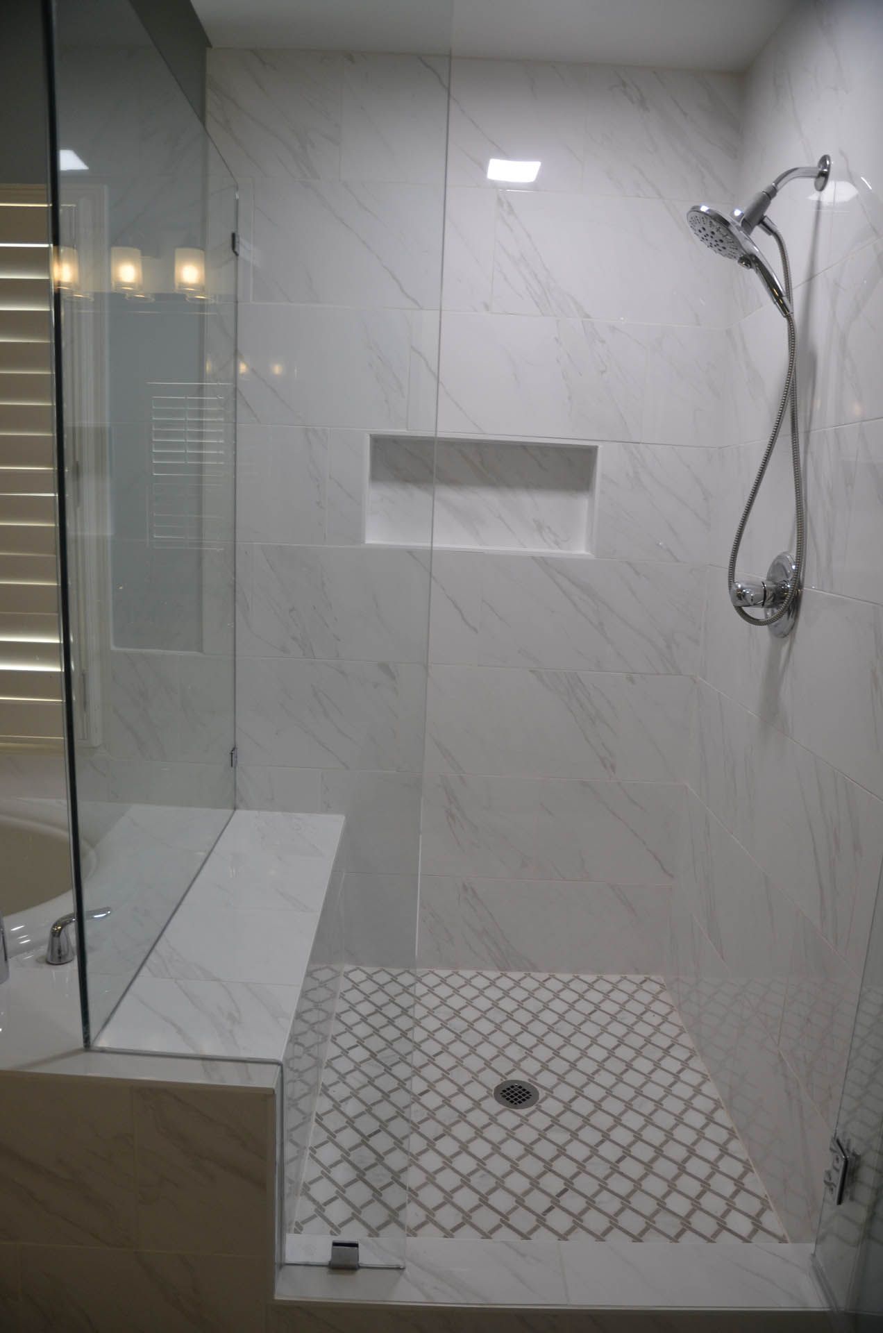 A modern, tiled shower with a glass door, built-in bench, and rain showerhead. The floor is mosaic tile, and the walls are white marble-like tiles.