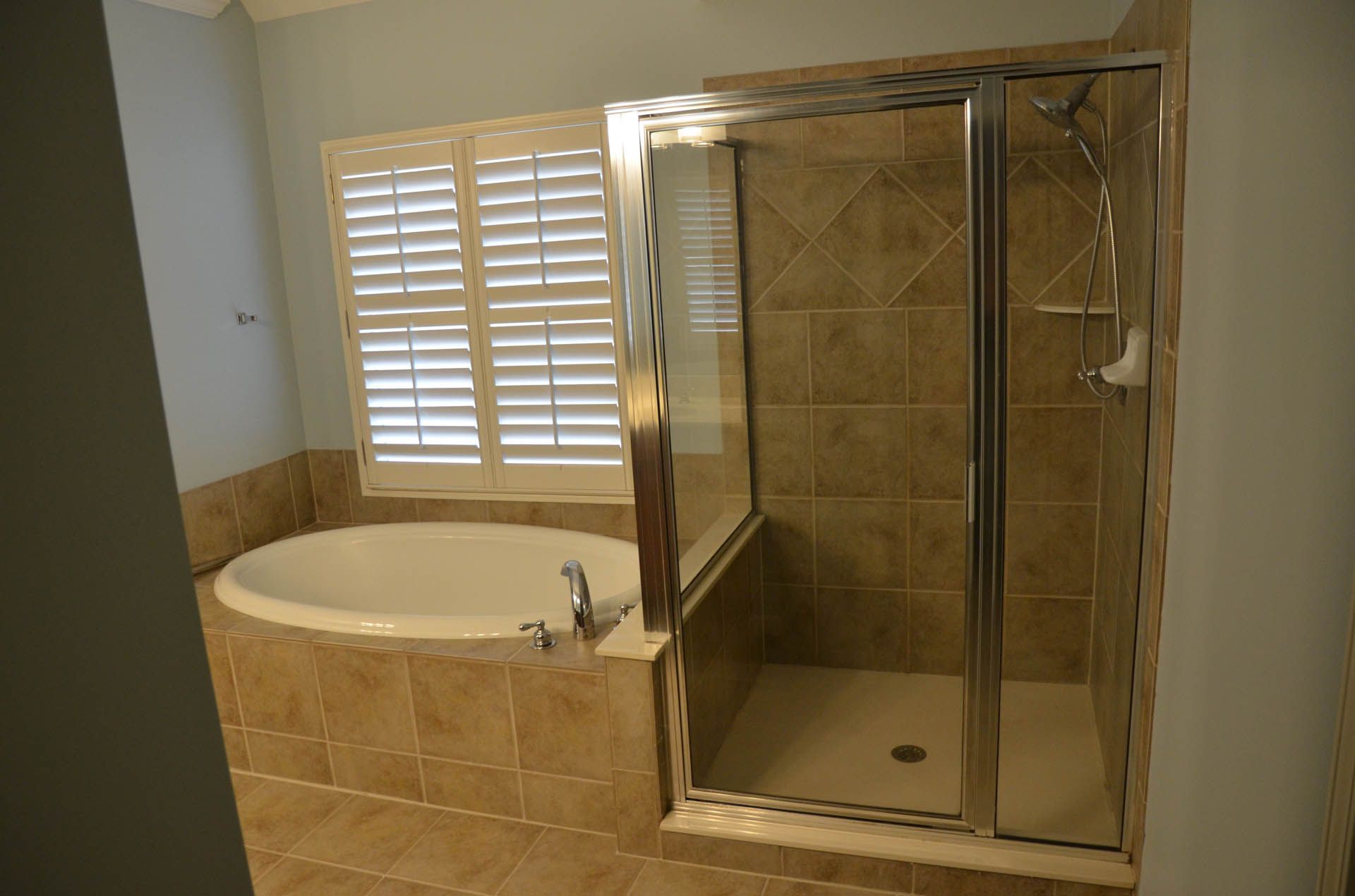 Bathroom with a white, oval-shaped tub, a glass shower, and a window with white shutters; tan tiles cover the floor and walls.