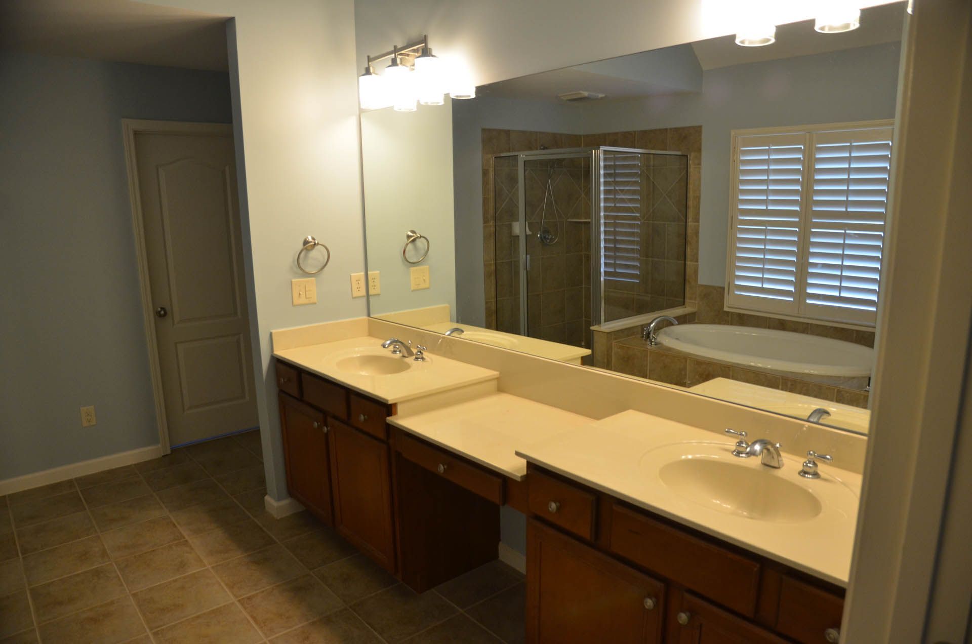 Spacious bathroom with dual sinks, large mirror, and a shower and bathtub visible in the background. Brown cabinets and tile floor.