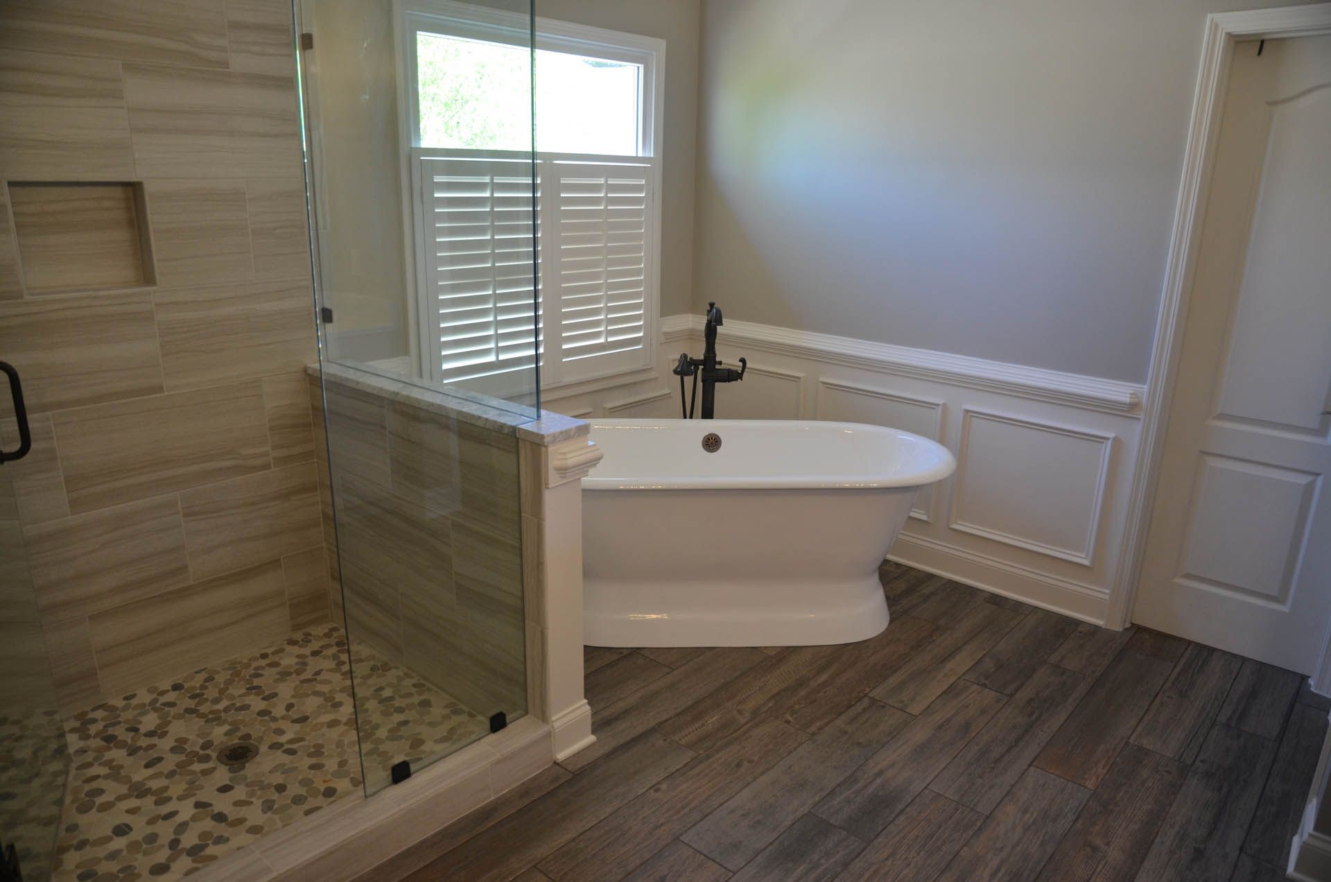 Spacious bathroom with a freestanding white bathtub, a glass-walled shower, and a window with white shutters.  Wood-look tile flooring.