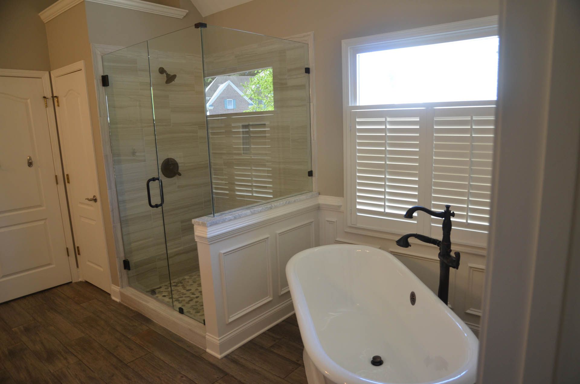 Bathroom with a glass-enclosed shower, freestanding tub, and a window with shutters. The floors are wood-look, and the walls are light-colored.