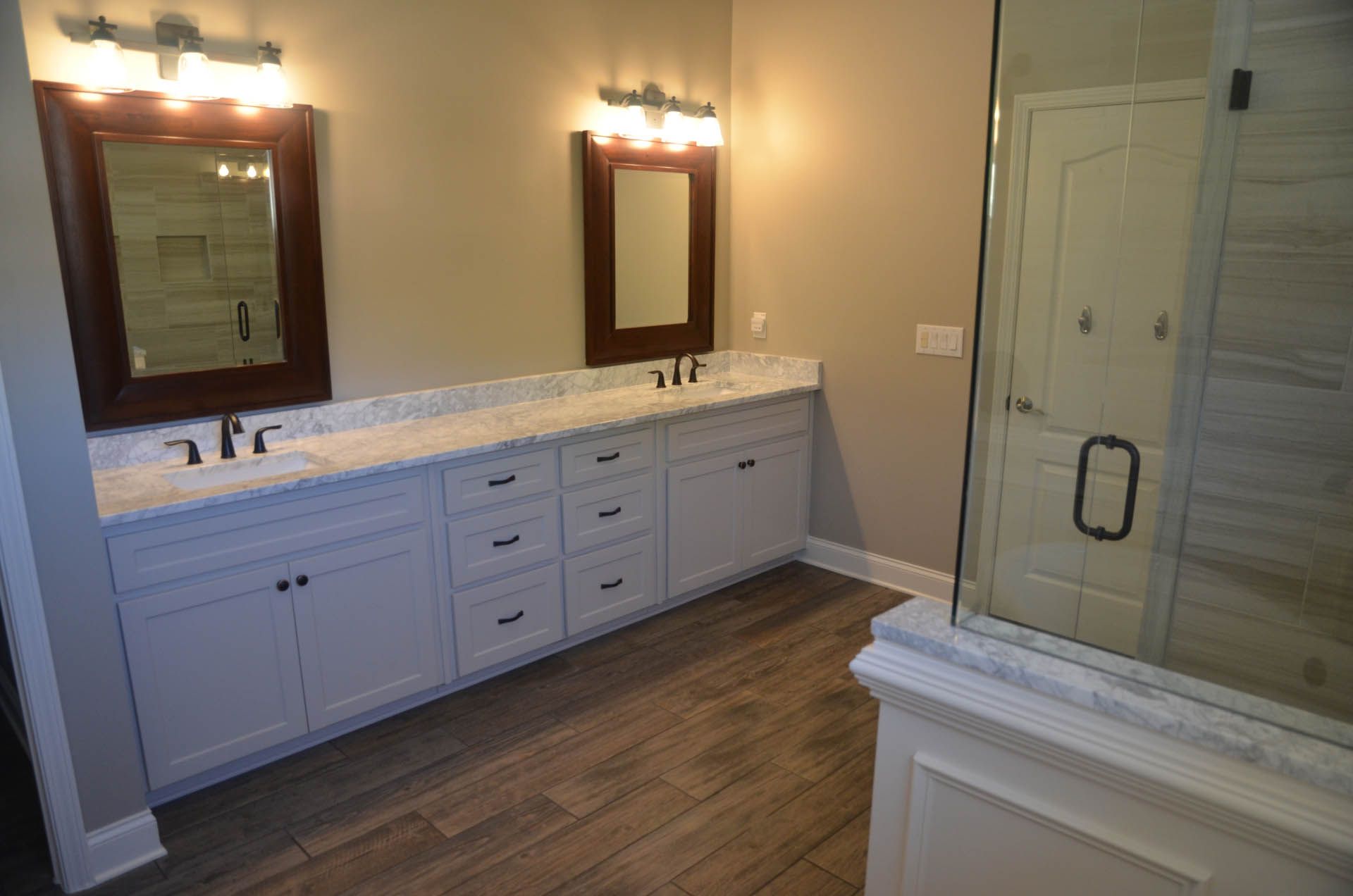 Bathroom with white cabinets, wood-look floor, two brown-framed mirrors, and a glass shower.