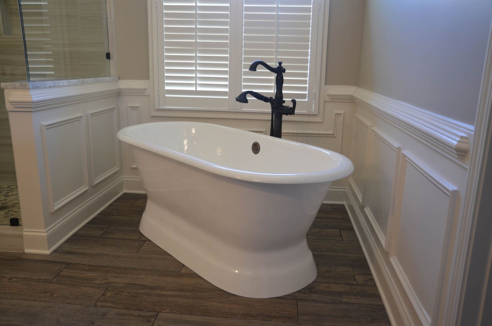 A white, freestanding bathtub with dark faucet in a bathroom with wood-look tile flooring and light-colored walls.