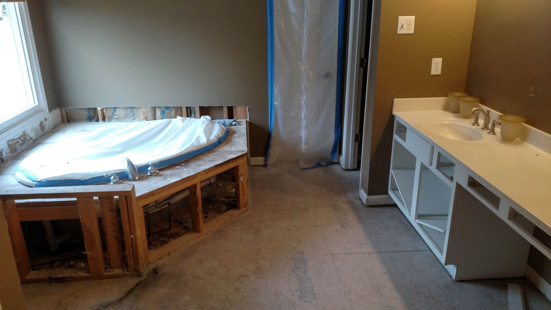 Bathroom under renovation. A built-in jacuzzi is near a window. A new vanity is in place, with no drawers or doors.