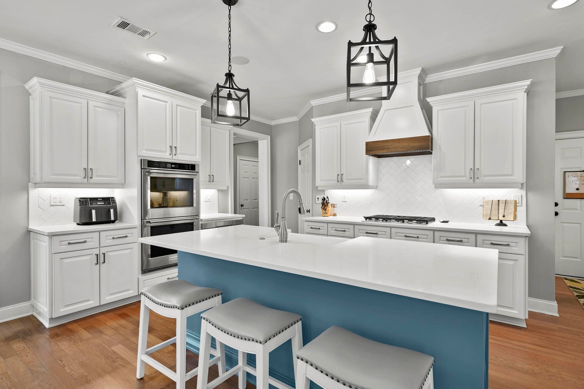A modern kitchen with white cabinets, a blue island, and wooden floors. Two black pendant lights hang above the island.