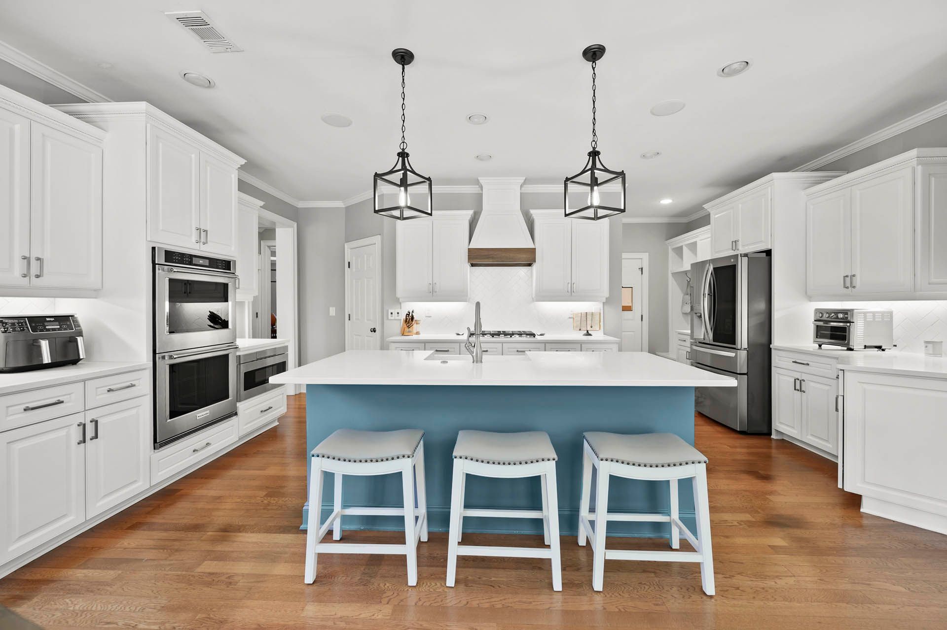 Modern kitchen with white cabinets, blue island, hardwood floors, and pendant lights. Three bar stools sit at the island.