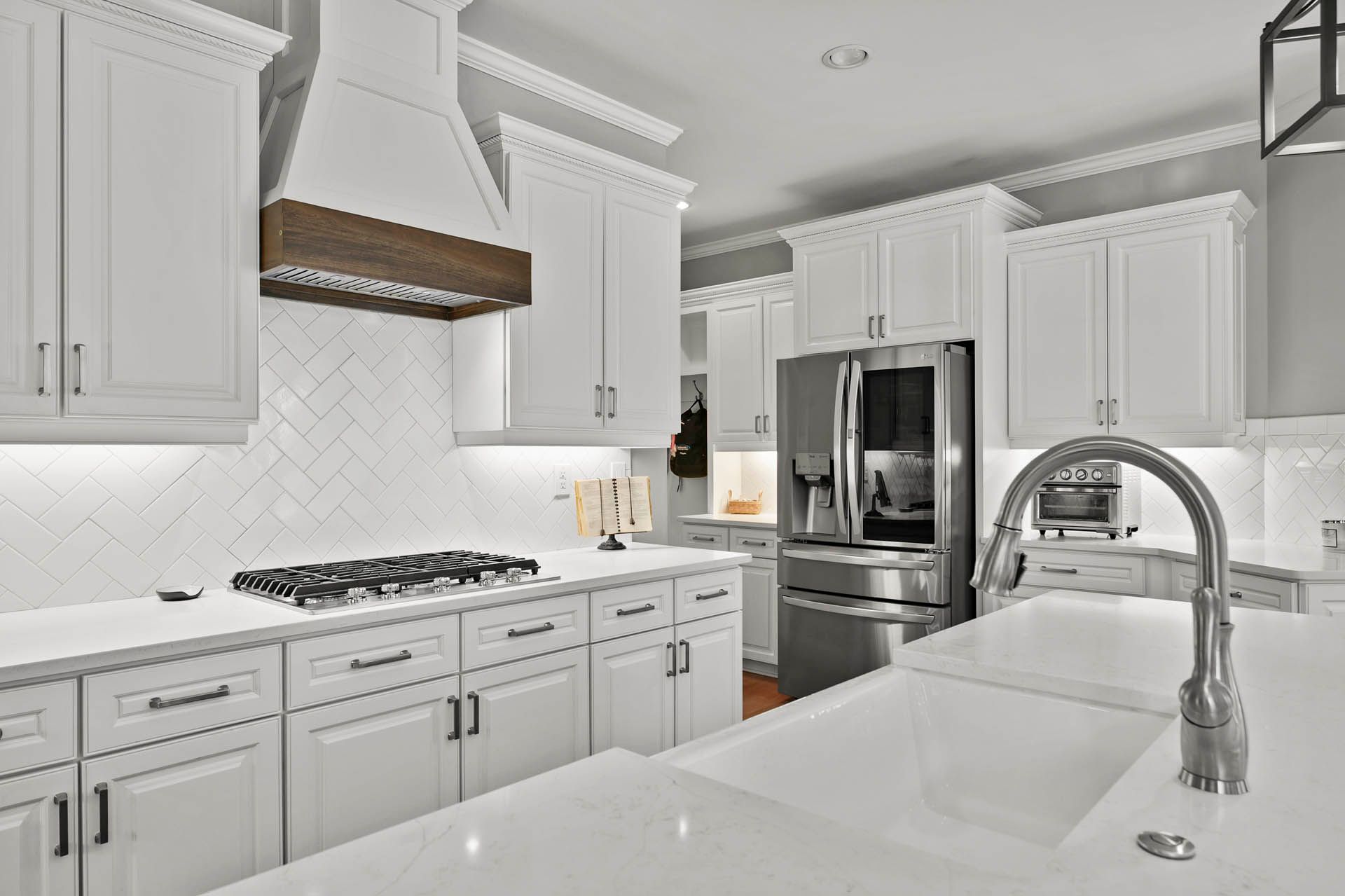 White kitchen with white cabinets, countertops, and a herringbone backsplash. Stainless steel appliances and a wooden range hood.