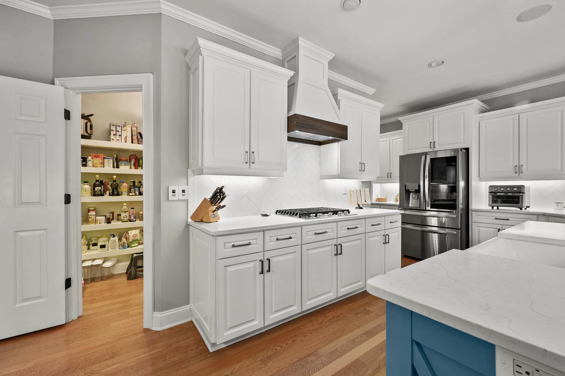 A modern kitchen with white cabinets, stainless steel appliances, and a blue island. A pantry door is open to reveal shelves stocked with food.