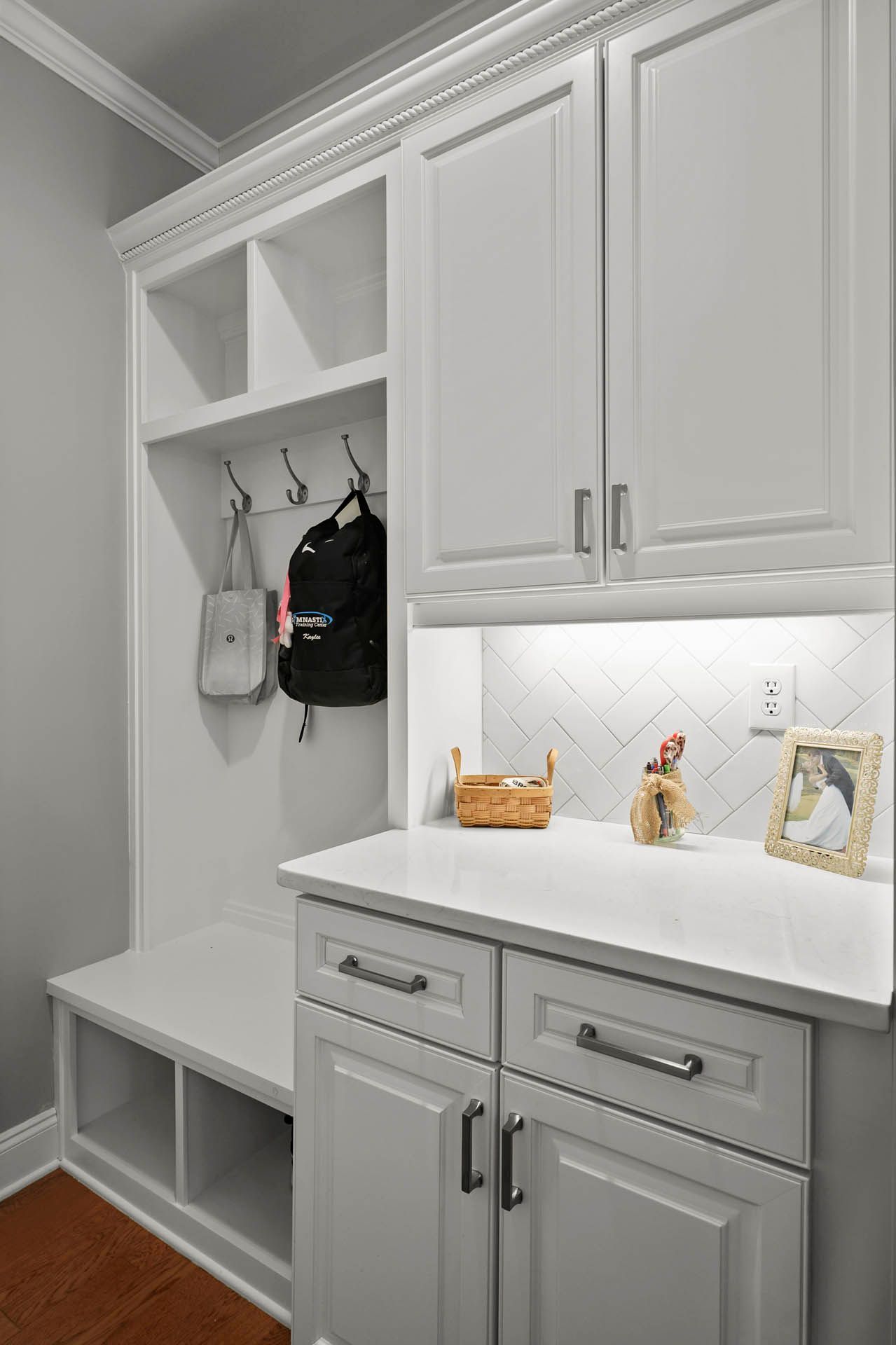 White built-in storage unit with open shelving, coat hooks, a bench, and cabinets. A white countertop has under-cabinet lighting and a herringbone tile backsplash.