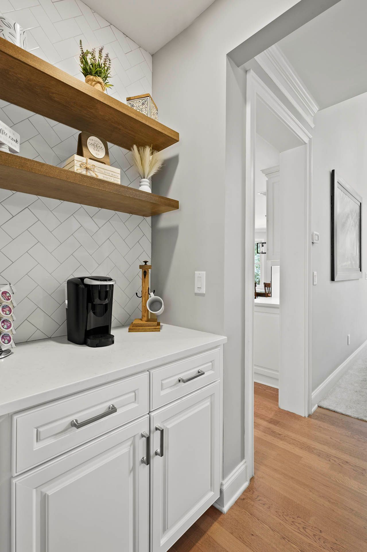 A coffee bar with white cabinets and a white countertop. Herringbone tiled backsplash, floating shelves with decor, and a doorway leading to another room.
