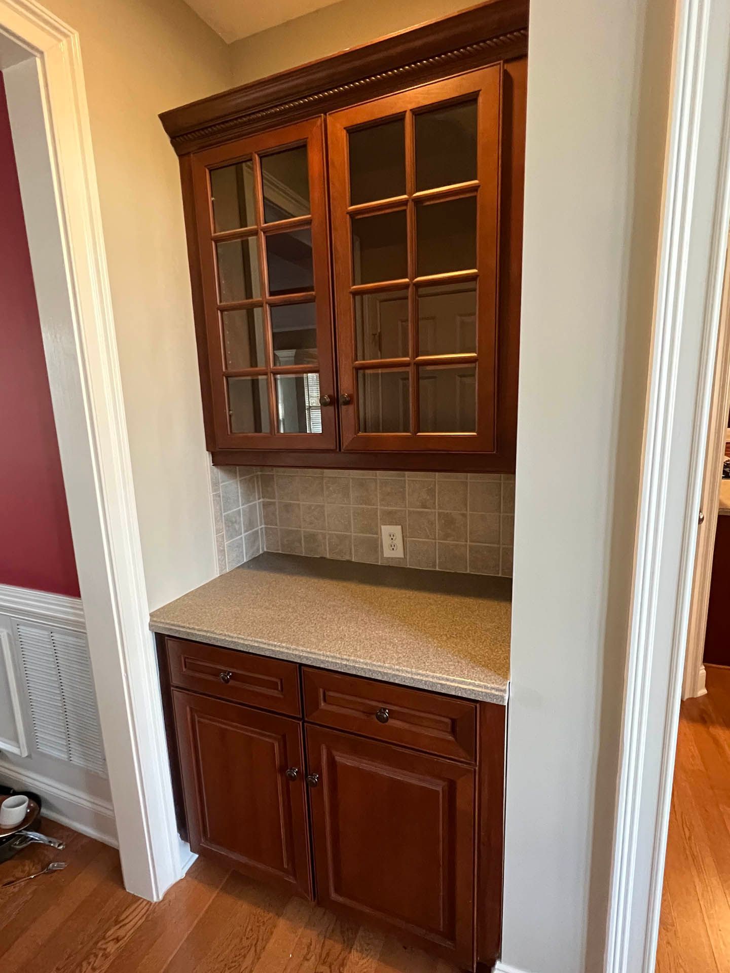 Built-in wooden cabinet with glass-paned doors, beige countertop, and two lower cabinets. Tile backsplash and an electrical outlet are visible.