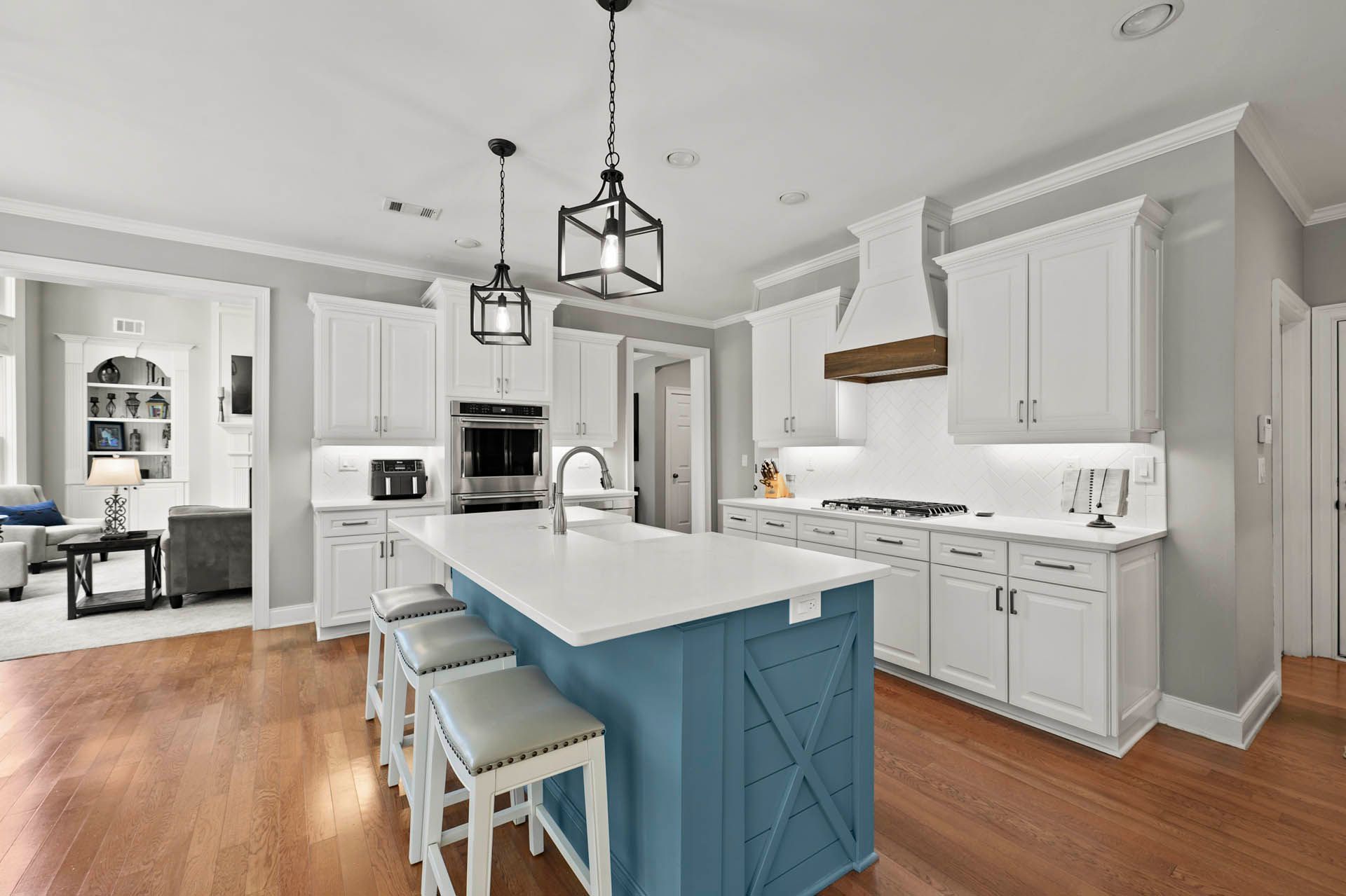 Spacious kitchen with white cabinetry, a blue island with seating, and wooden floors. A living area is visible in the background.