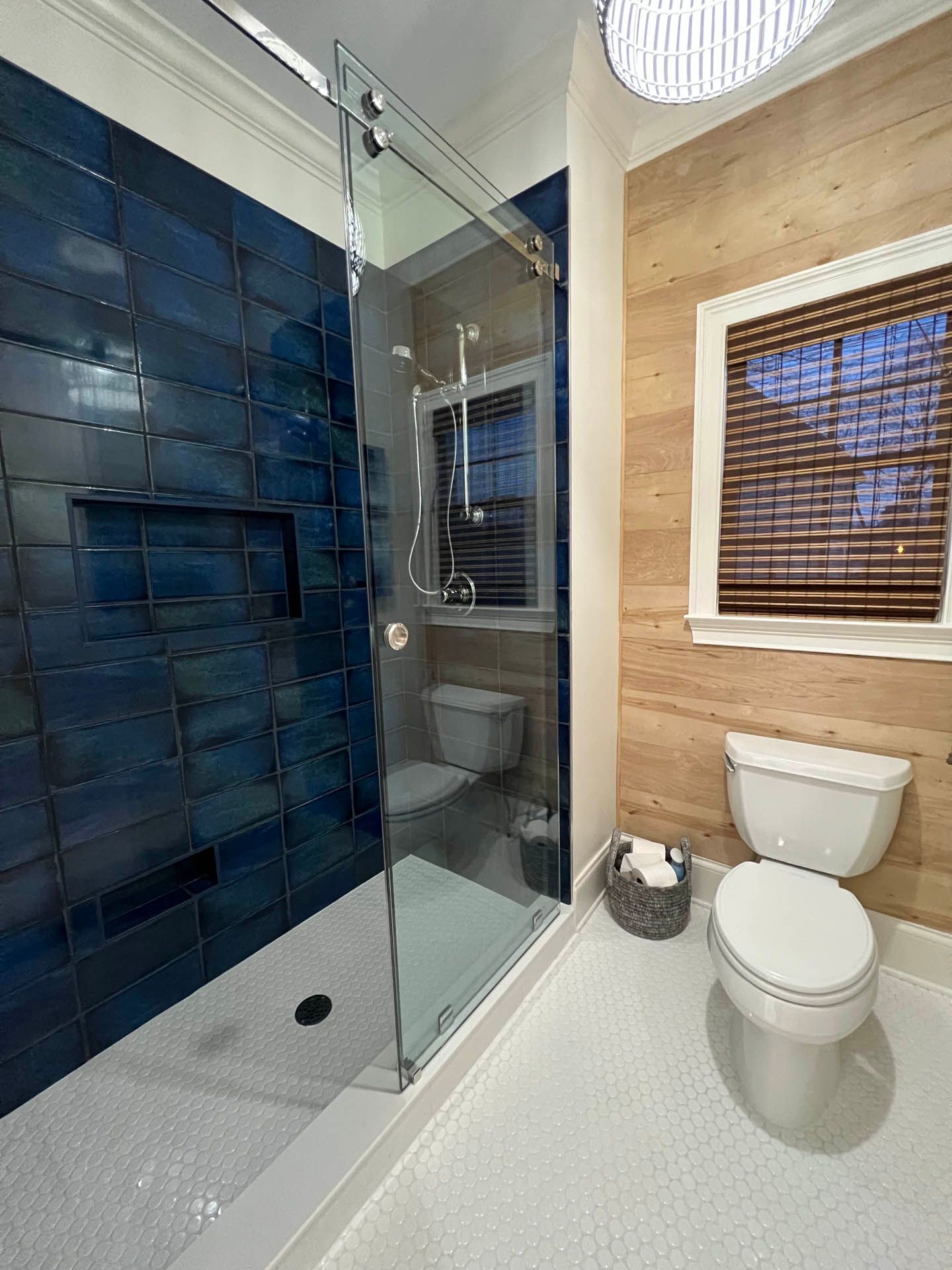 Bathroom with blue tiled shower, sliding glass door, and wooden plank wall with a toilet and window.