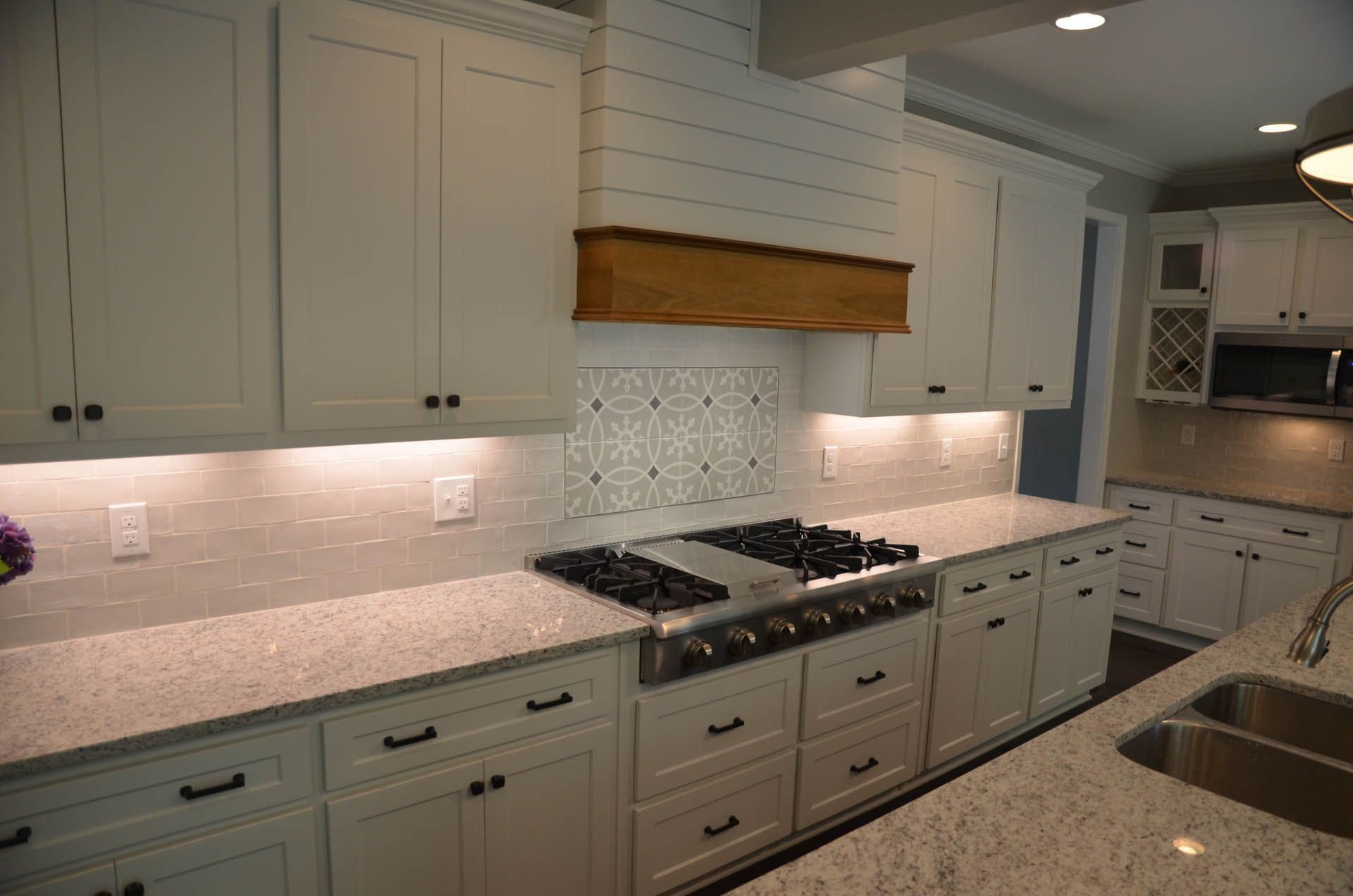 White kitchen with granite countertops, white cabinets, and a gas stovetop. The backsplash features a mosaic pattern, and there's a wooden range hood.